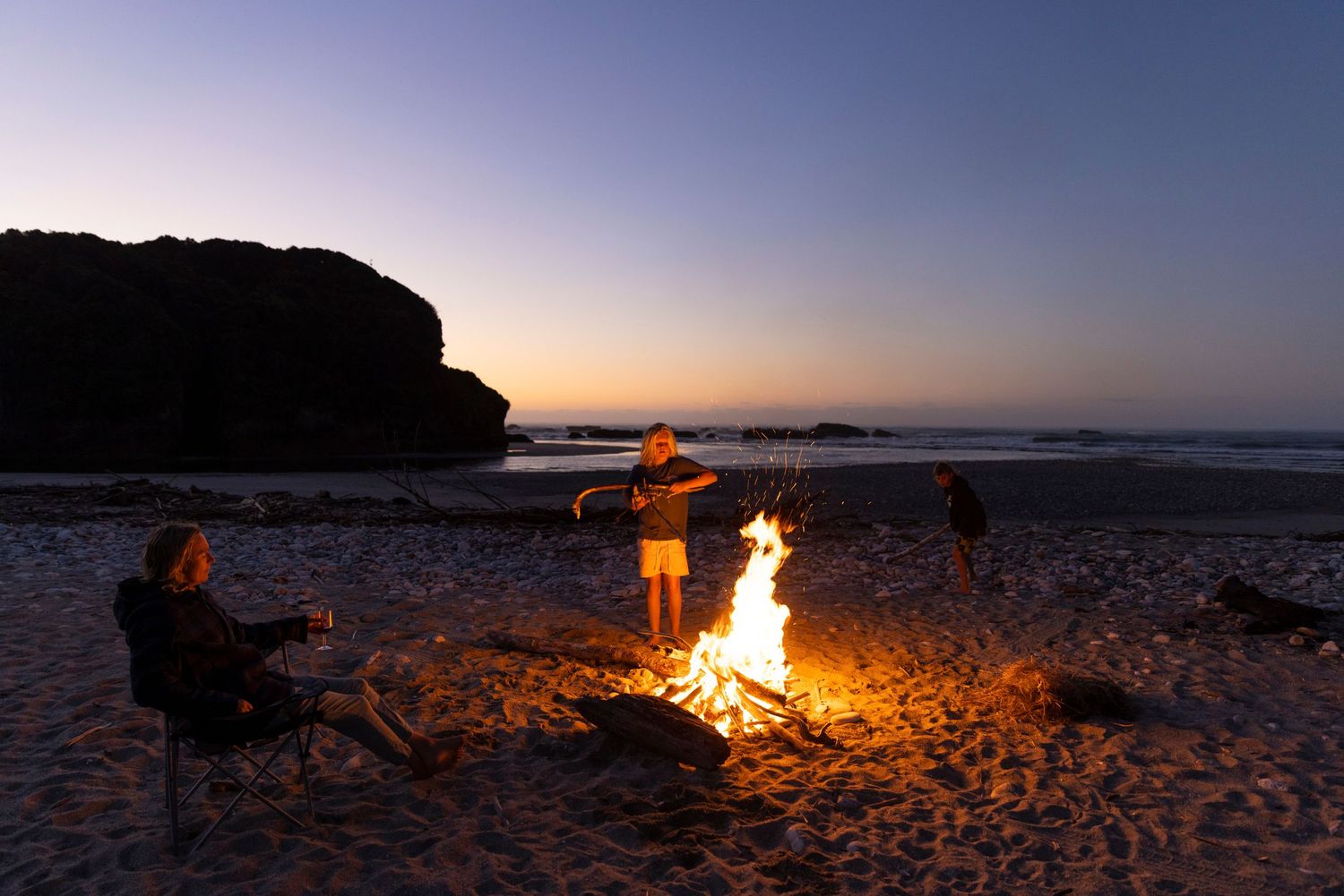 Beach bonfire scene at sunset with silhouettes against orange sky.