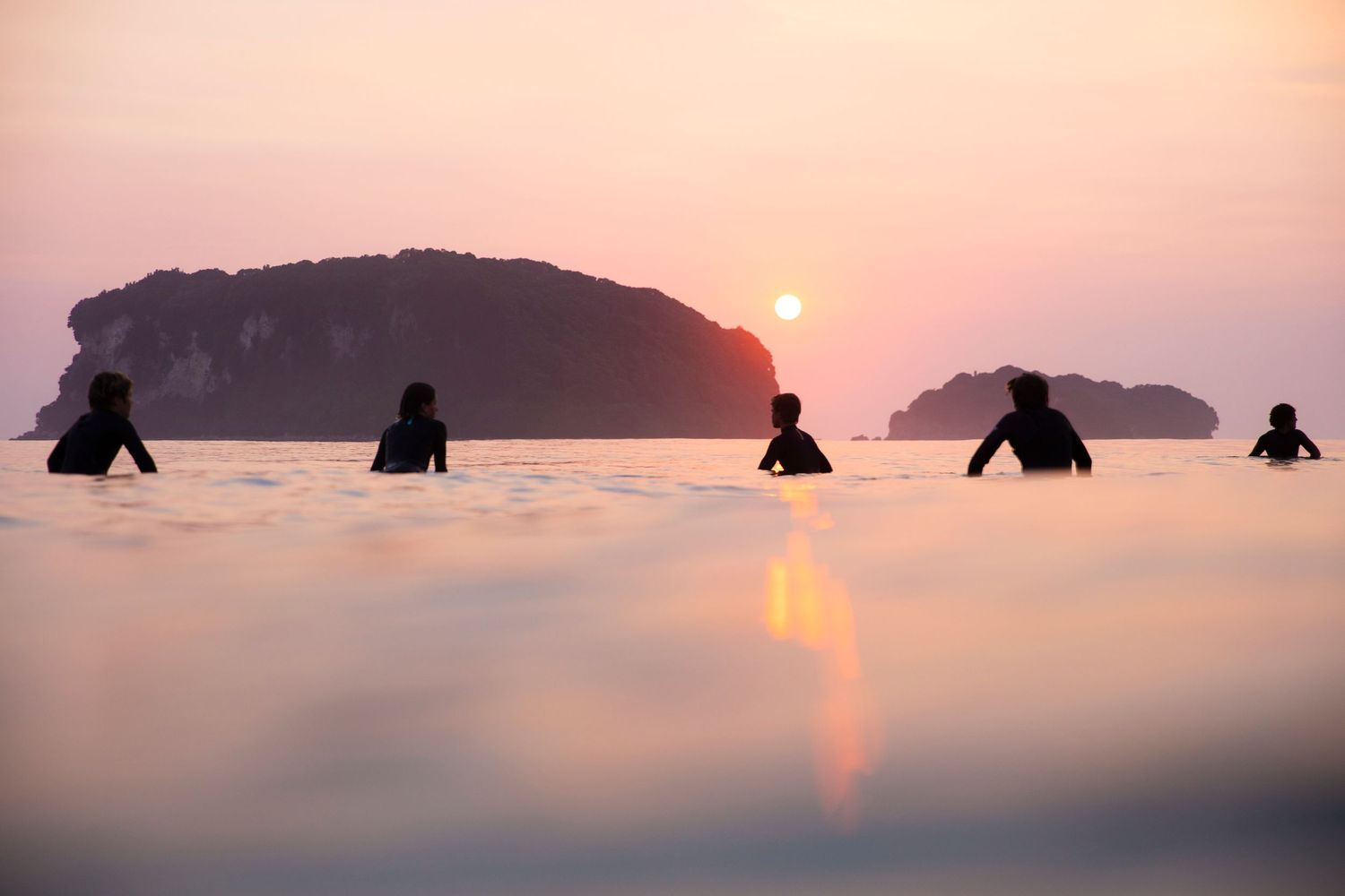 Pink sunset over calm waters with silhouettes of surfers in the distance.