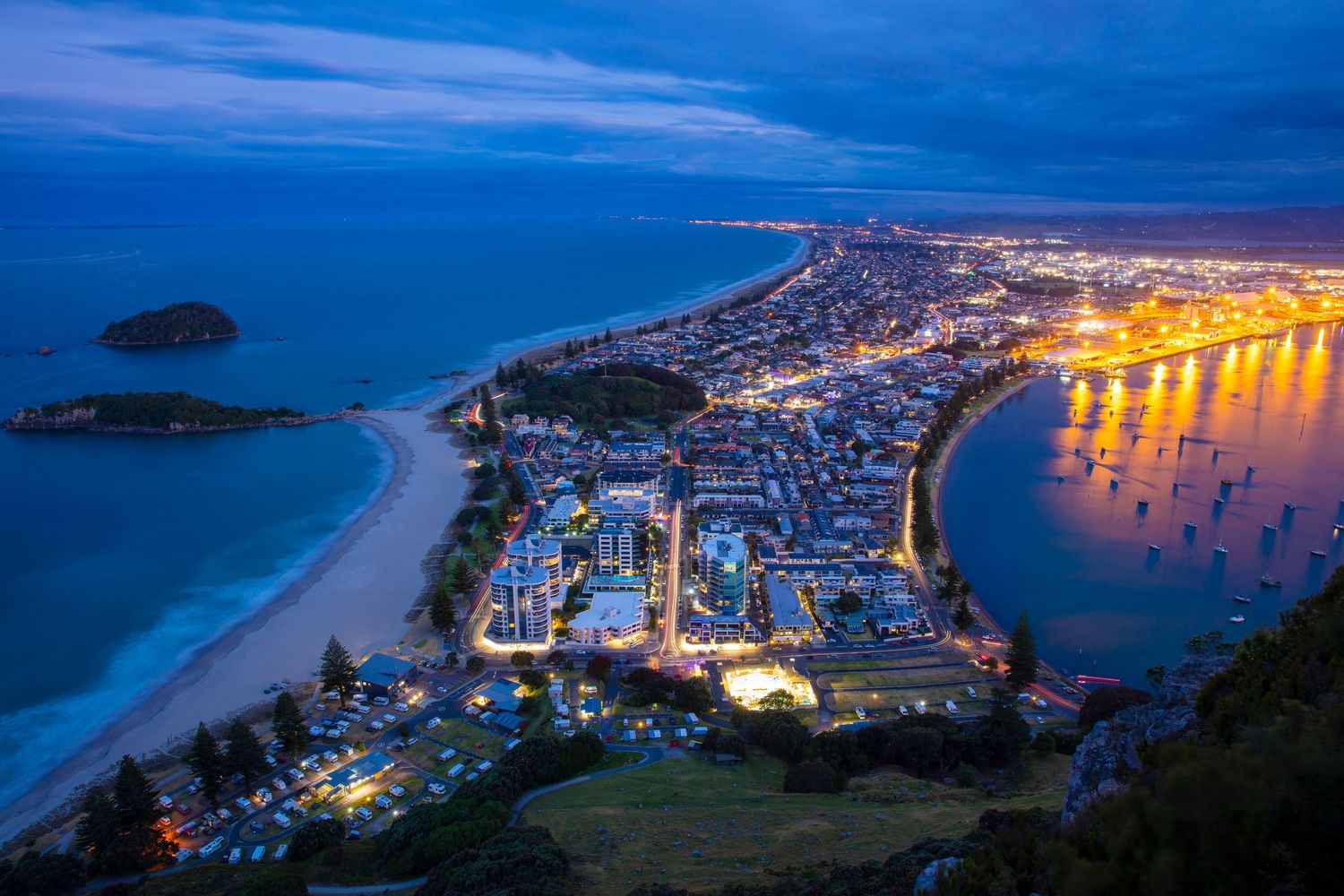 Panoramic nighttime cityscape of Mount Maunganui – a coastal town with twinkling lights reflecting on the harbor.