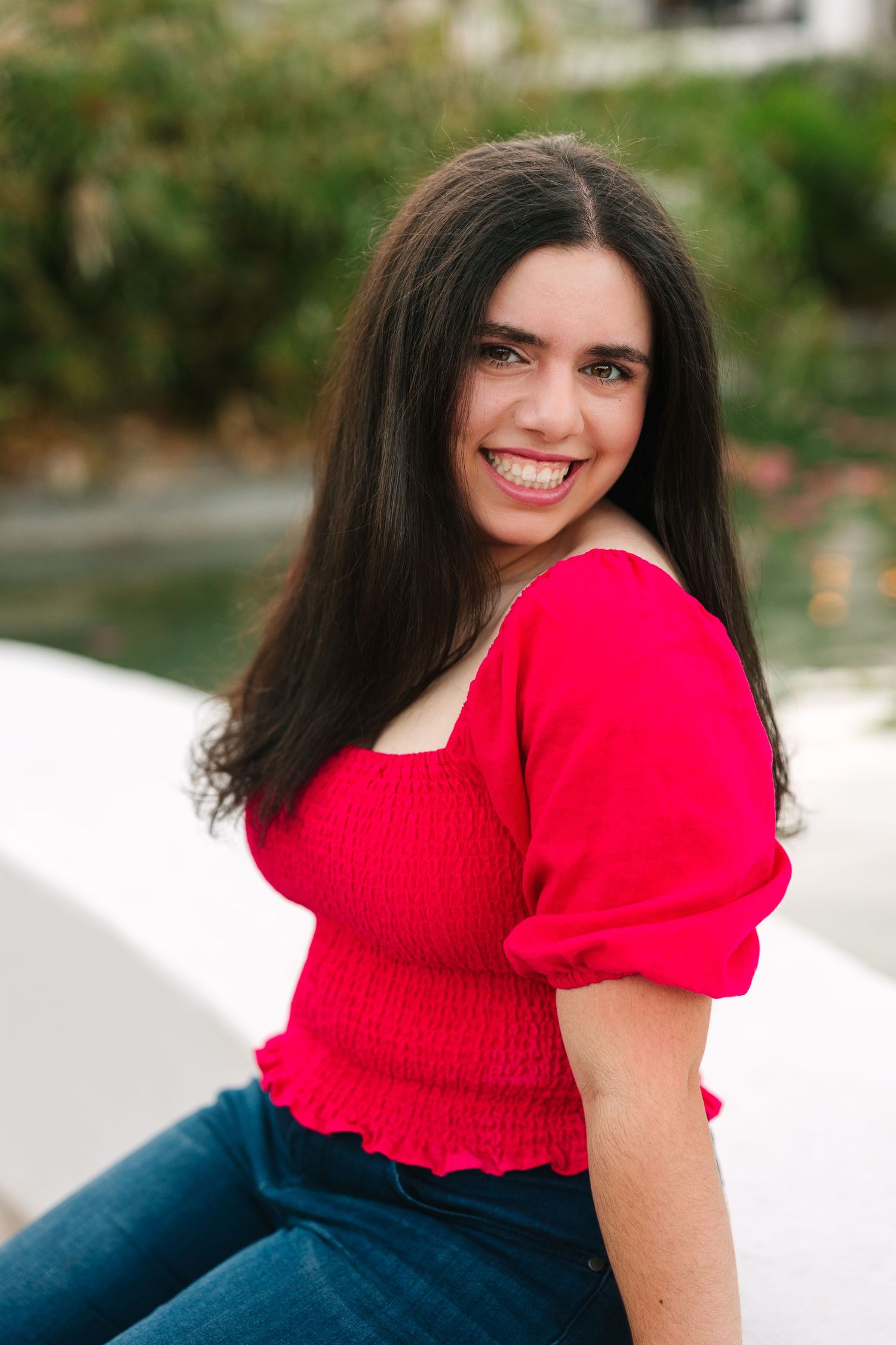 A smiling woman in a bright red smocked top and jeans poses outdoors against a blurred natural background.