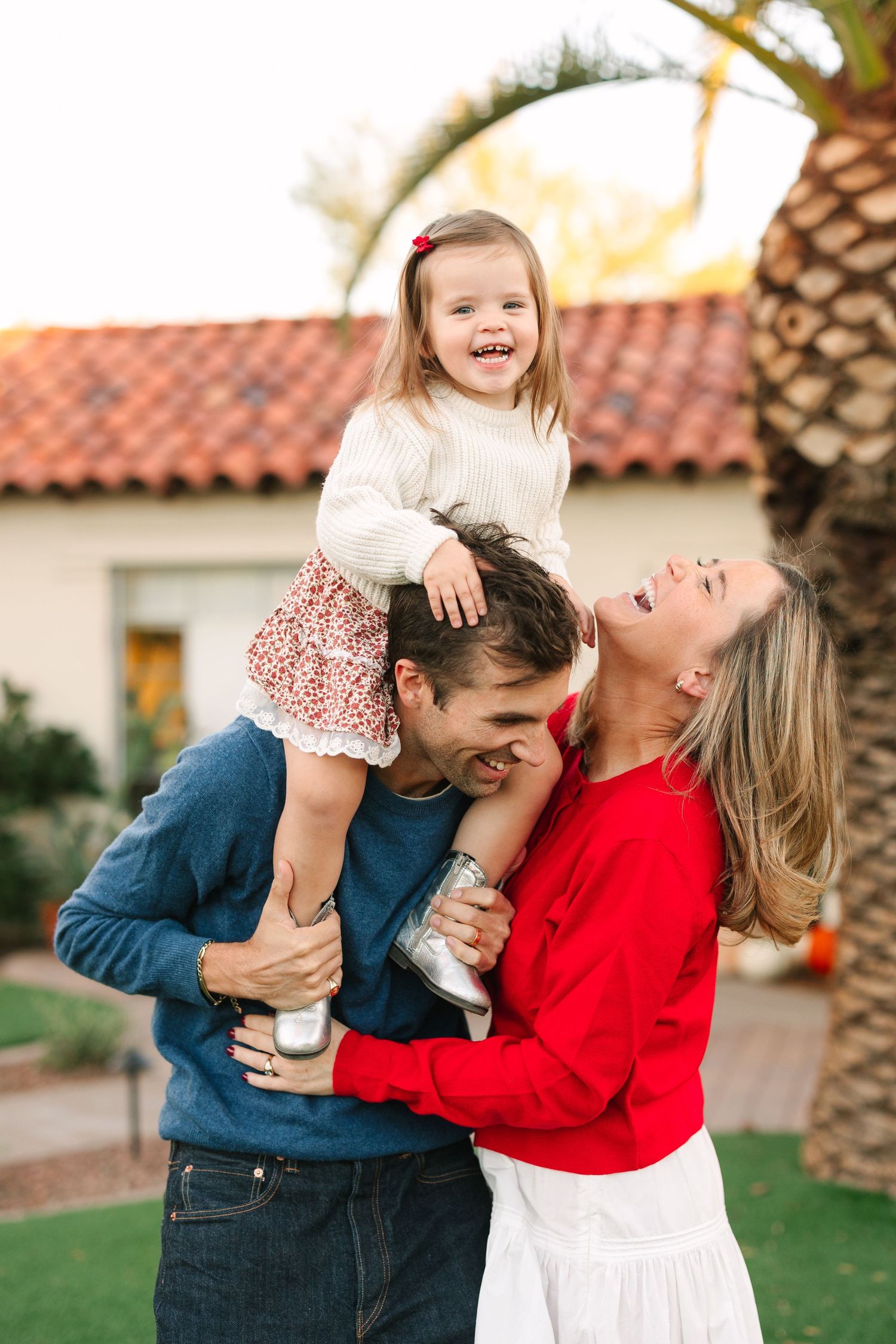 A joyful family shares a playful moment together outdoors on their lawn with Spanish tile roofing and palm trees in background.