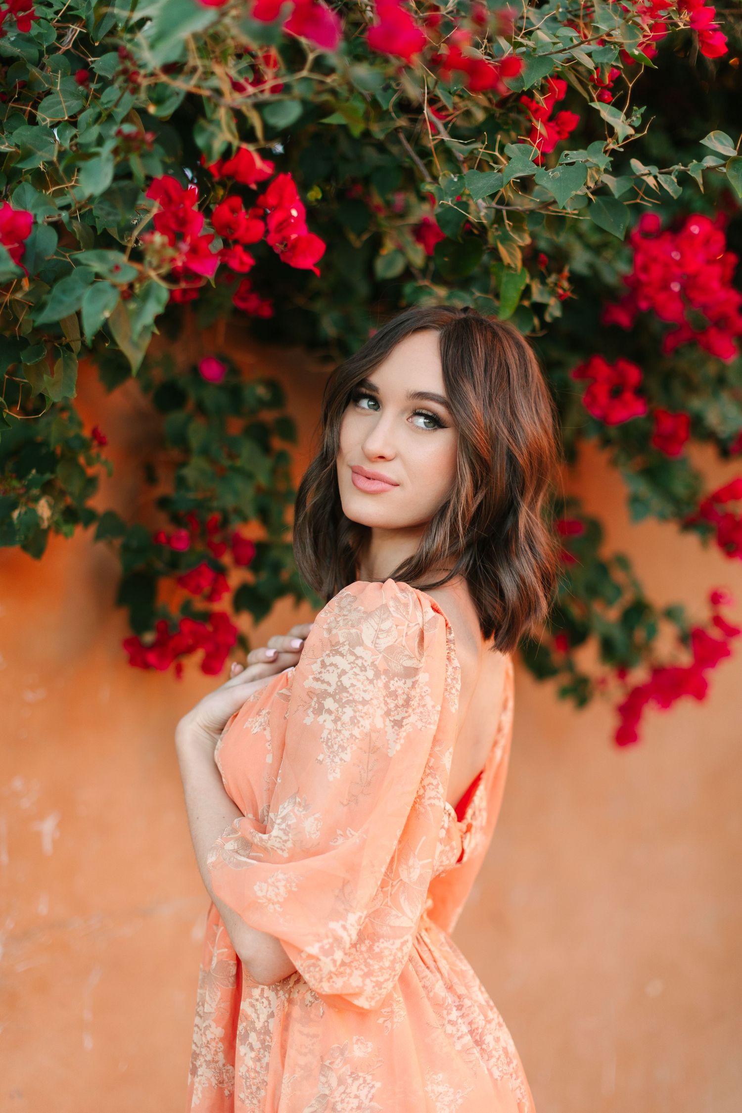 A person in a peach-colored dress poses beneath vibrant red flowers against a coral-colored wall.