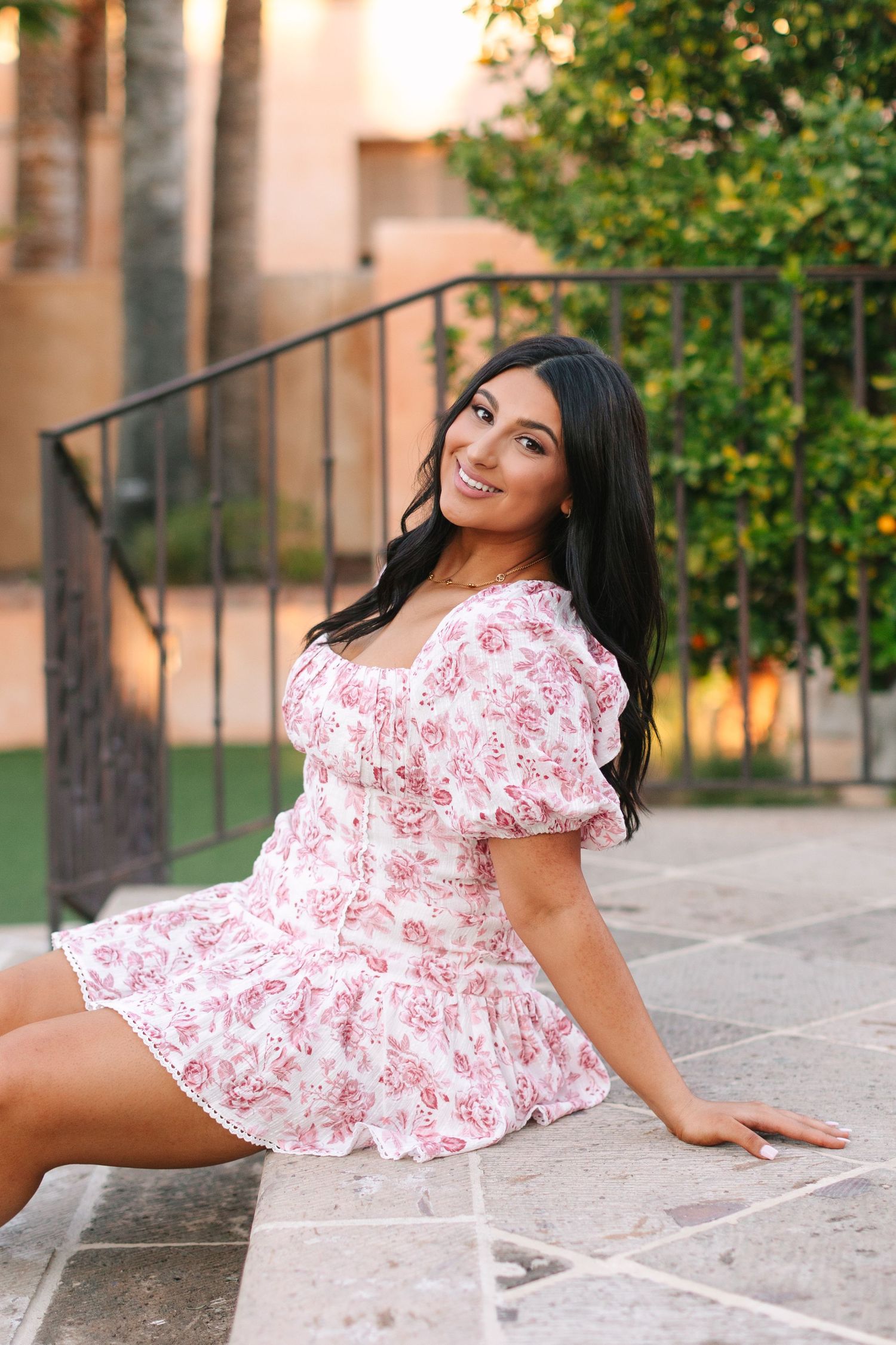 Woman in pink and white floral dress sits on stone paving during golden hour with greenery in background.