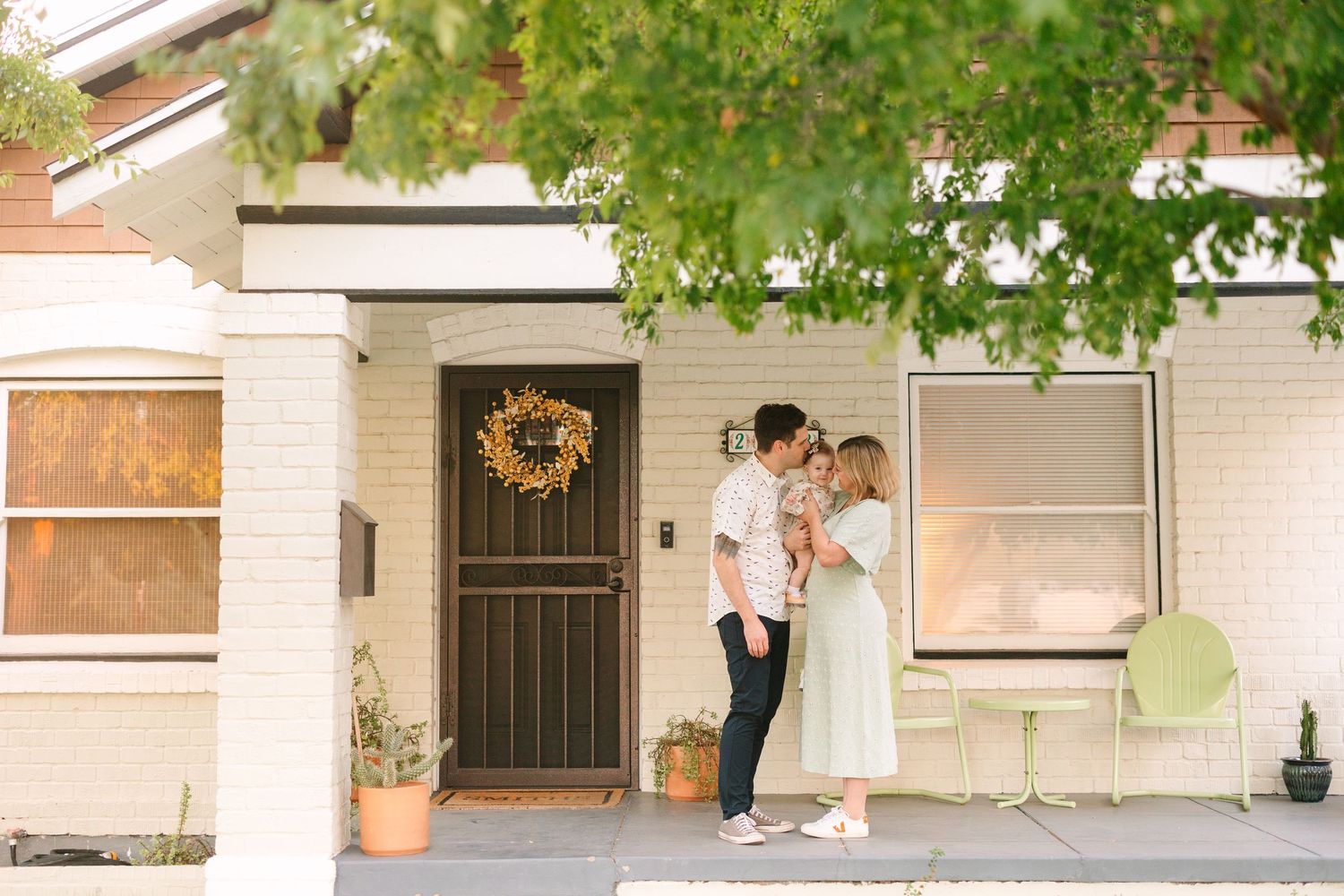 A couple stands together on a charming front porch with a decorative wreath hanging on the black security door.