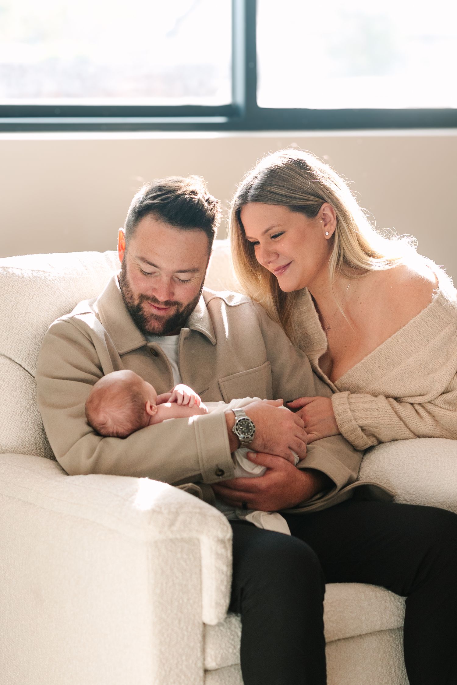 Loving parents sitting on white couch sharing tender moment with newborn baby in sunlit room.