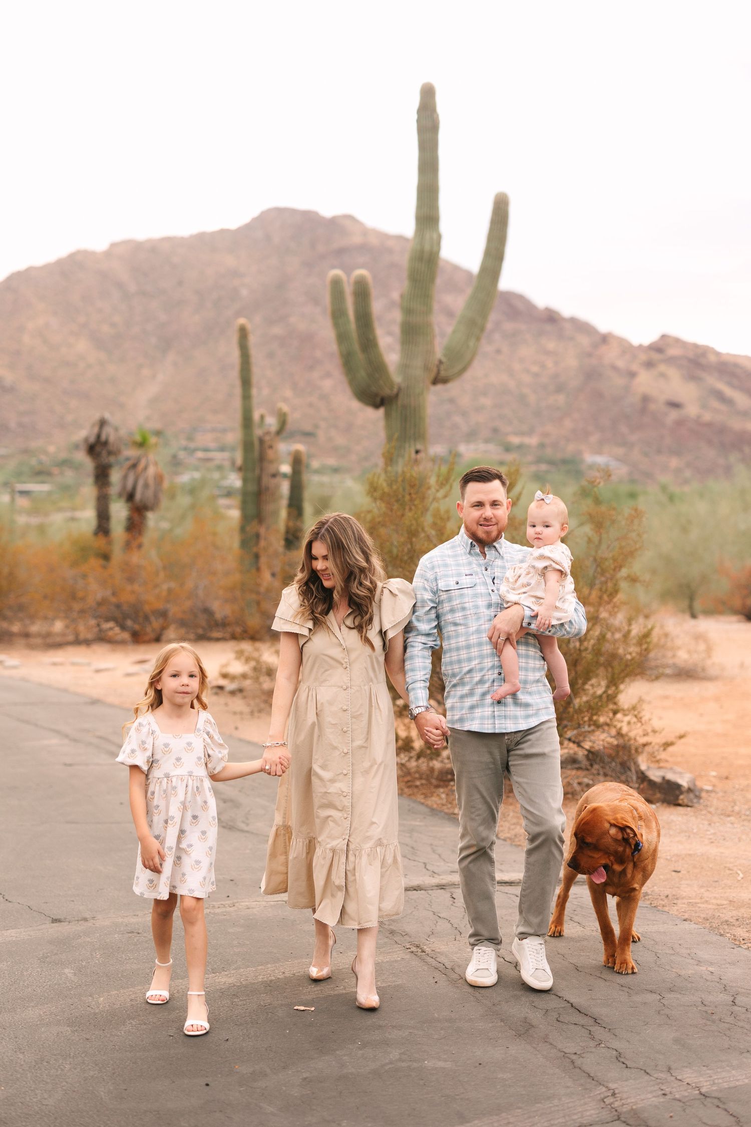 A family walks together on a desert path with their dog as towering saguaro cacti and mountains create a scenic backdrop.
