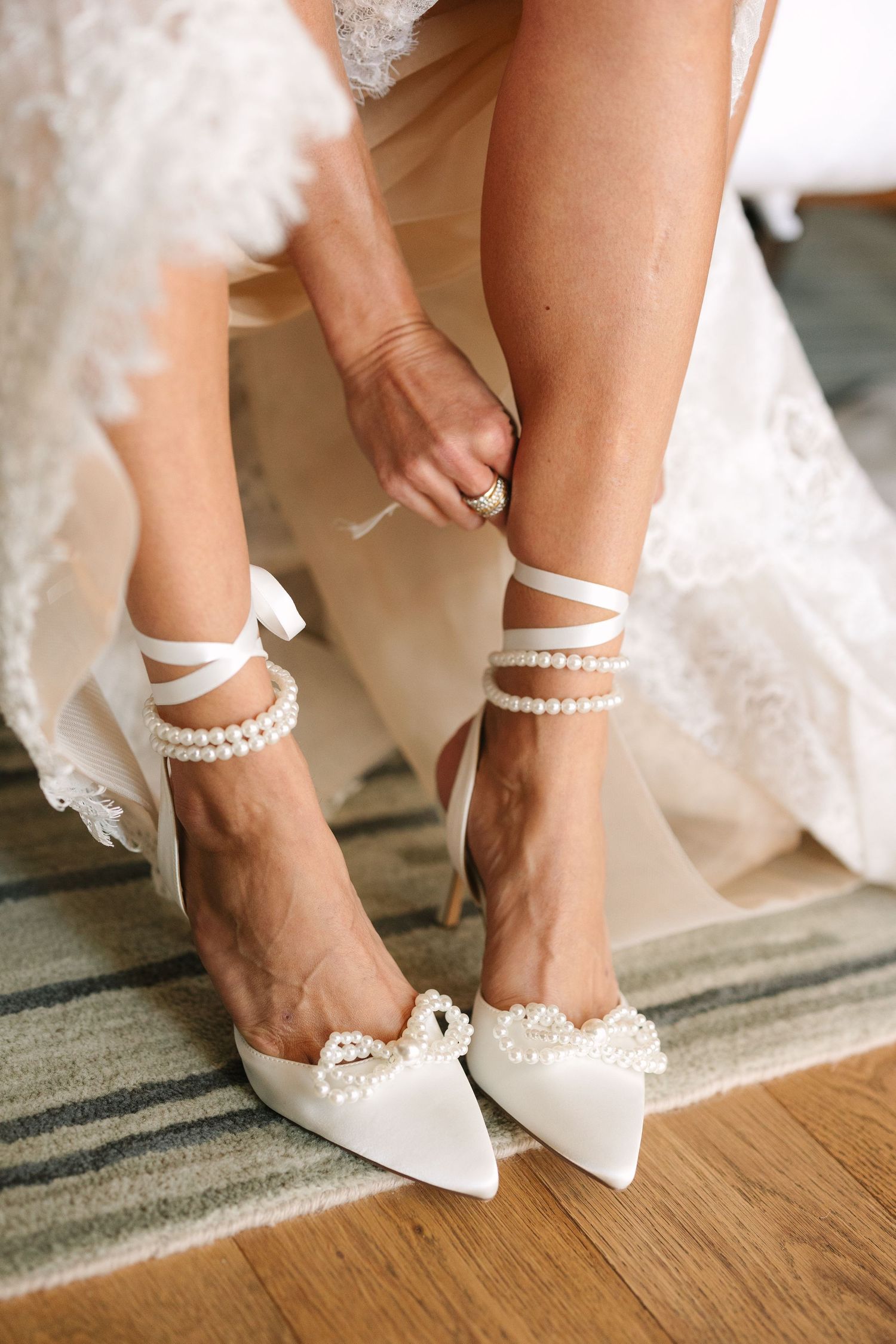 Elegant bridal shoes with pearl ankle straps and pointed toes being secured on hardwood floor.