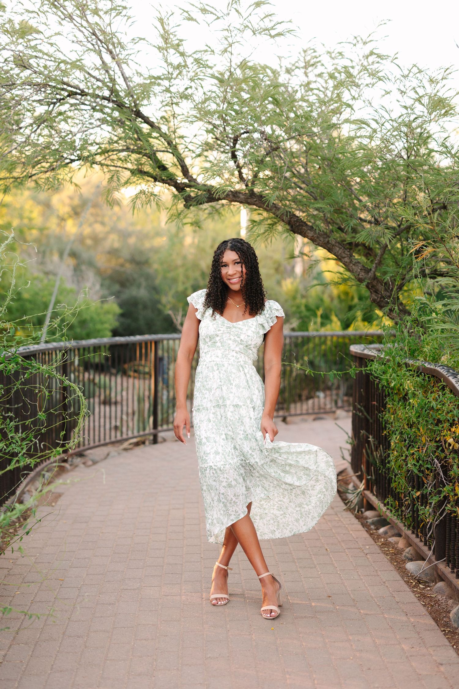 A white summery floral dress flows in the breeze along a garden pathway lined with lush trees and railings.