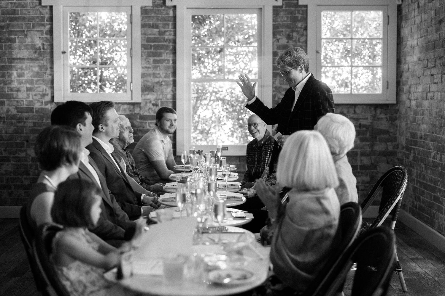 A black and white photo of dinner gathering with guests seated at a long table in a rustic brick-walled room.