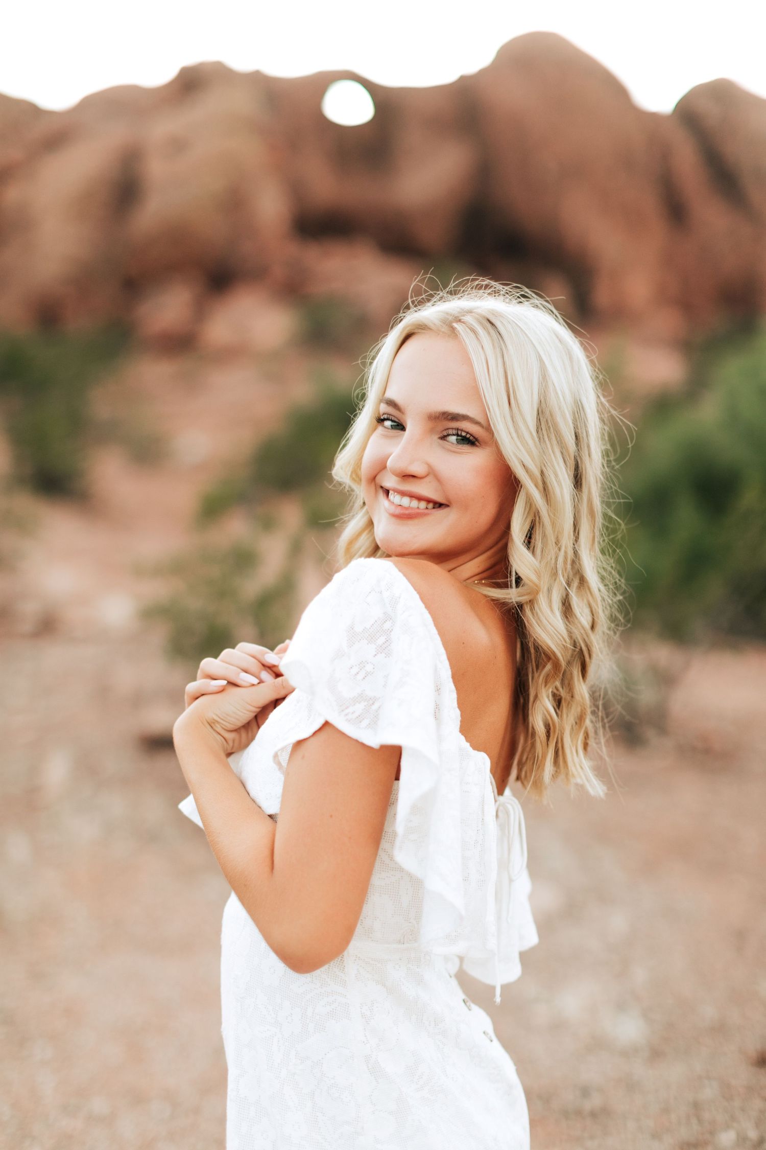 A woman in a white ruffled dress smiles while posing against red rock formations in a desert landscape.