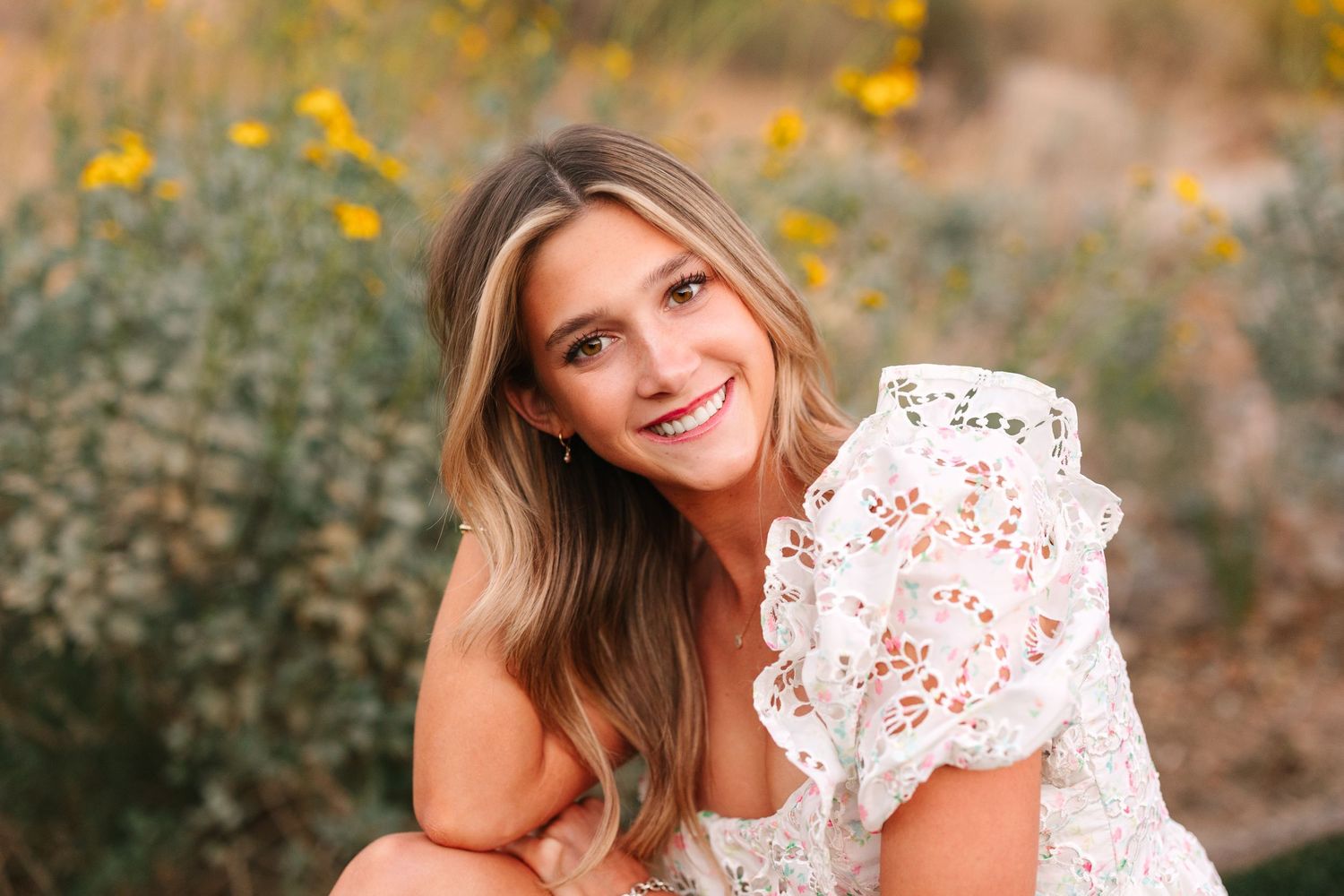 A person in a white lace dress poses outdoors with yellow wildflowers in a natural setting during golden hour.