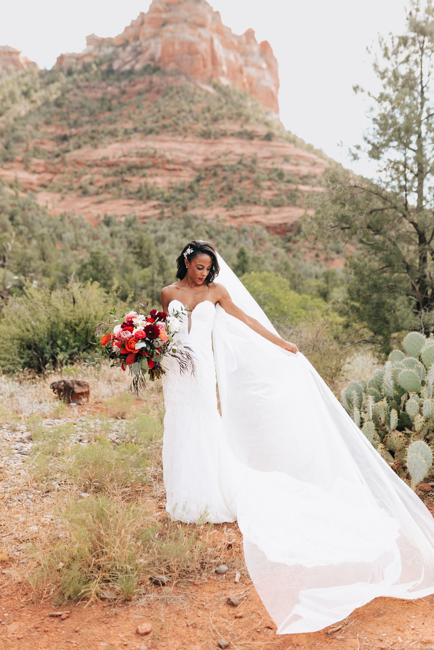 A bride in a flowing white gown poses dramatically against desert red rocks and cacti.
