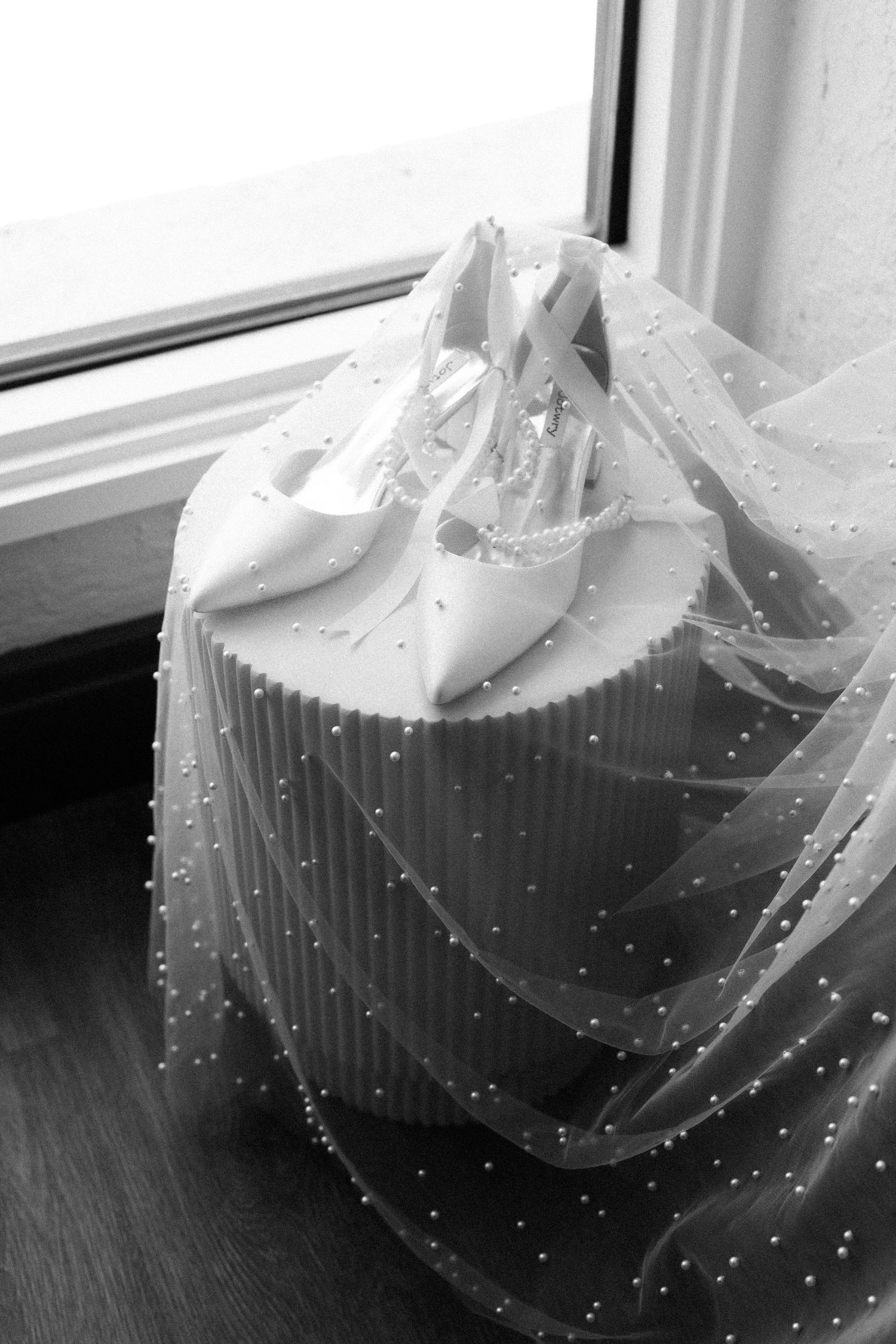 White bridal shoes resting on delicate beaded veil near window in black and white photography.