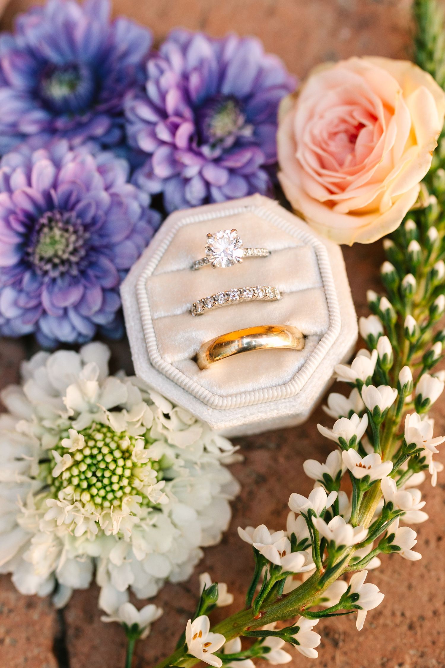 Wedding rings displayed on velvet box with purple and peach flowers.