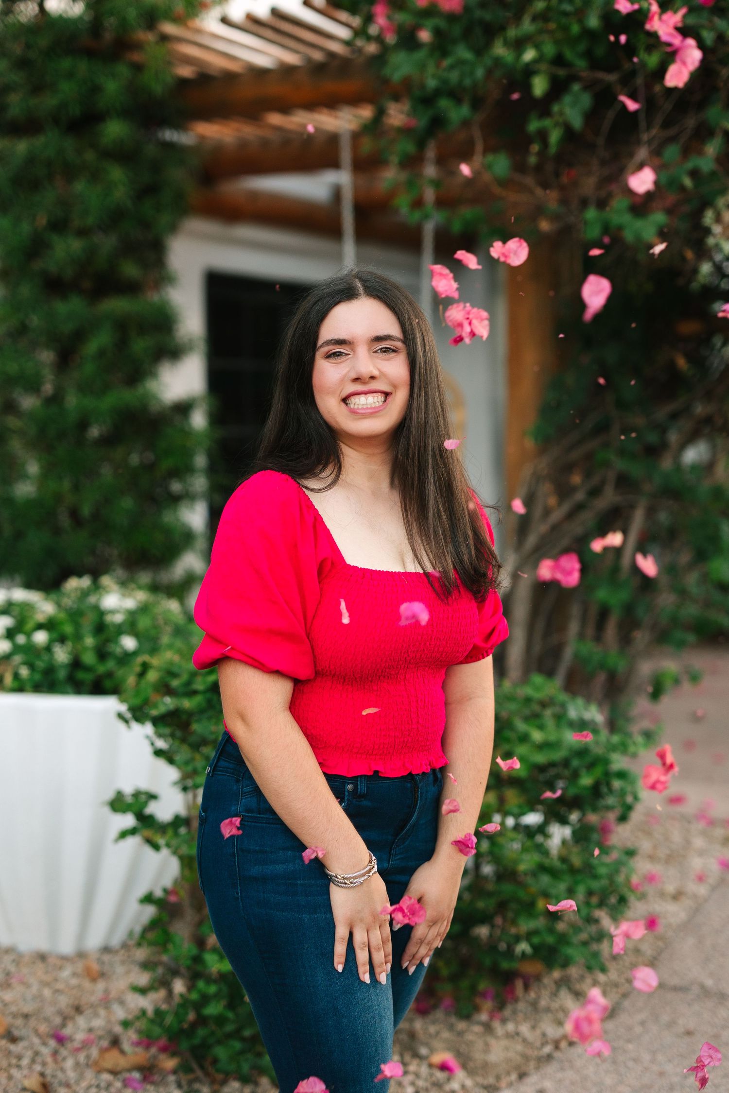 Someone wearing a bright red top and dark jeans poses in a garden setting with pink flower petals falling in the foreground.