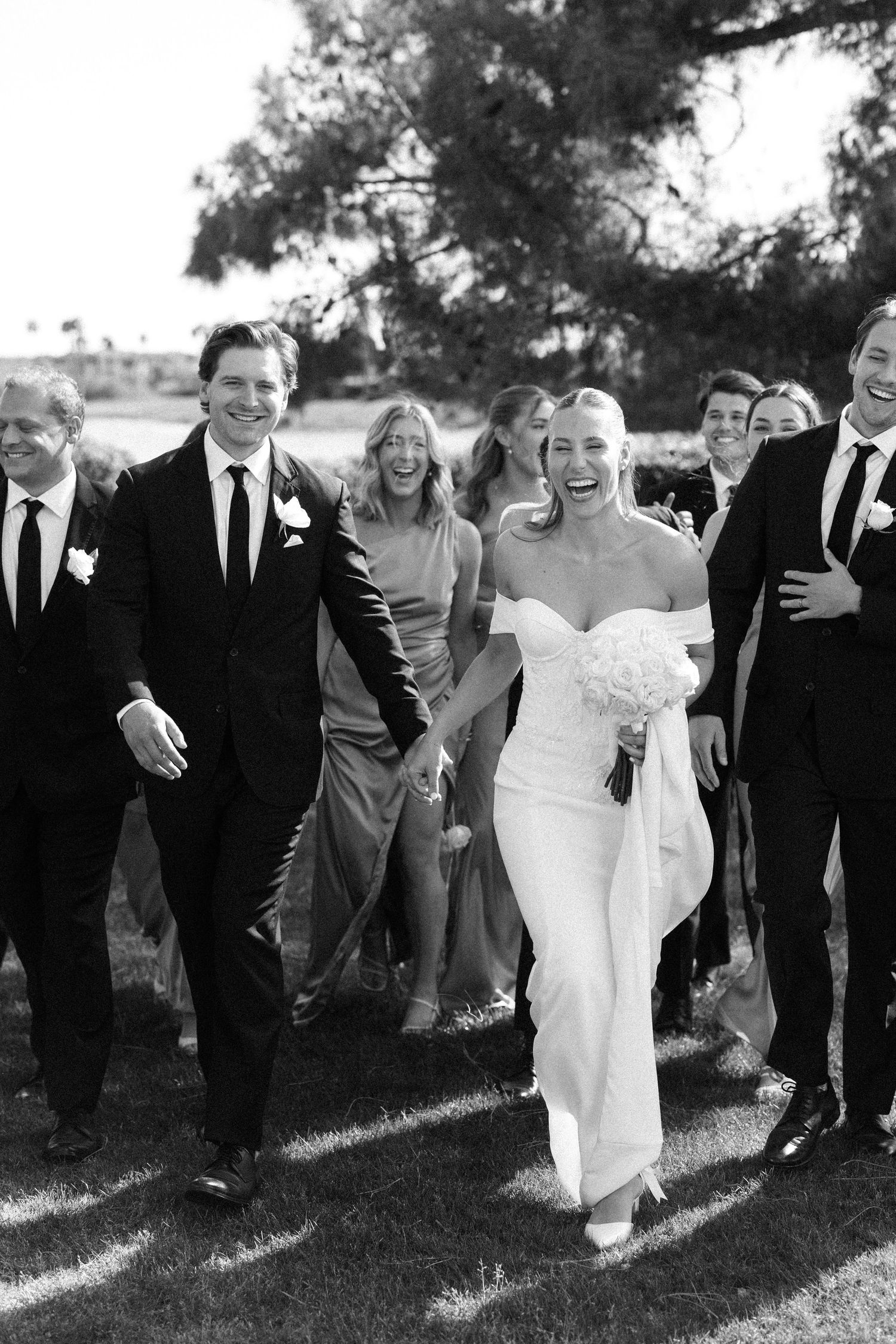 A joyful wedding party in formal attire laughs and walks together outdoors in black and white photography.