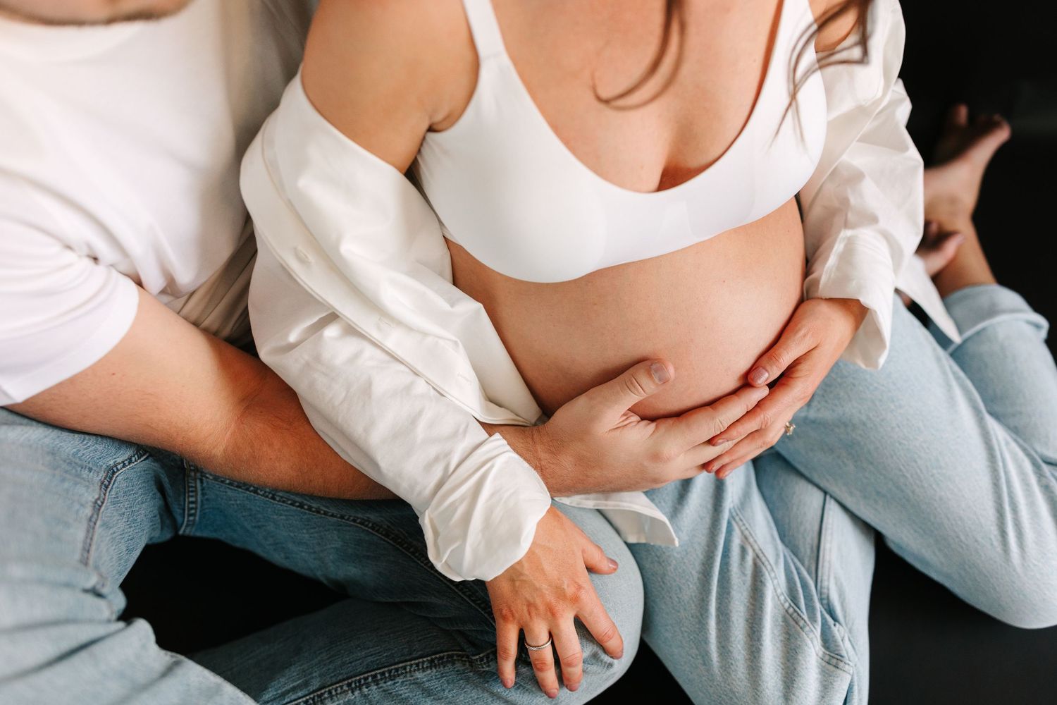 Close up view of hands tenderly embracing a pregnant belly while wearing a white sports bra and light jeans.
