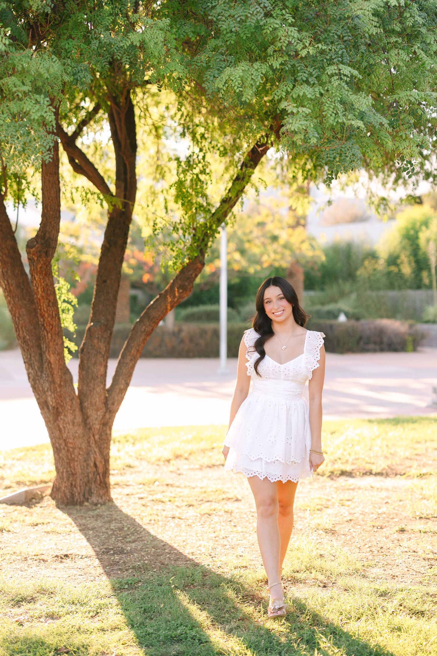 Woman in white summer dress poses outdoors under tree branches in warm golden sunlight at sunset.