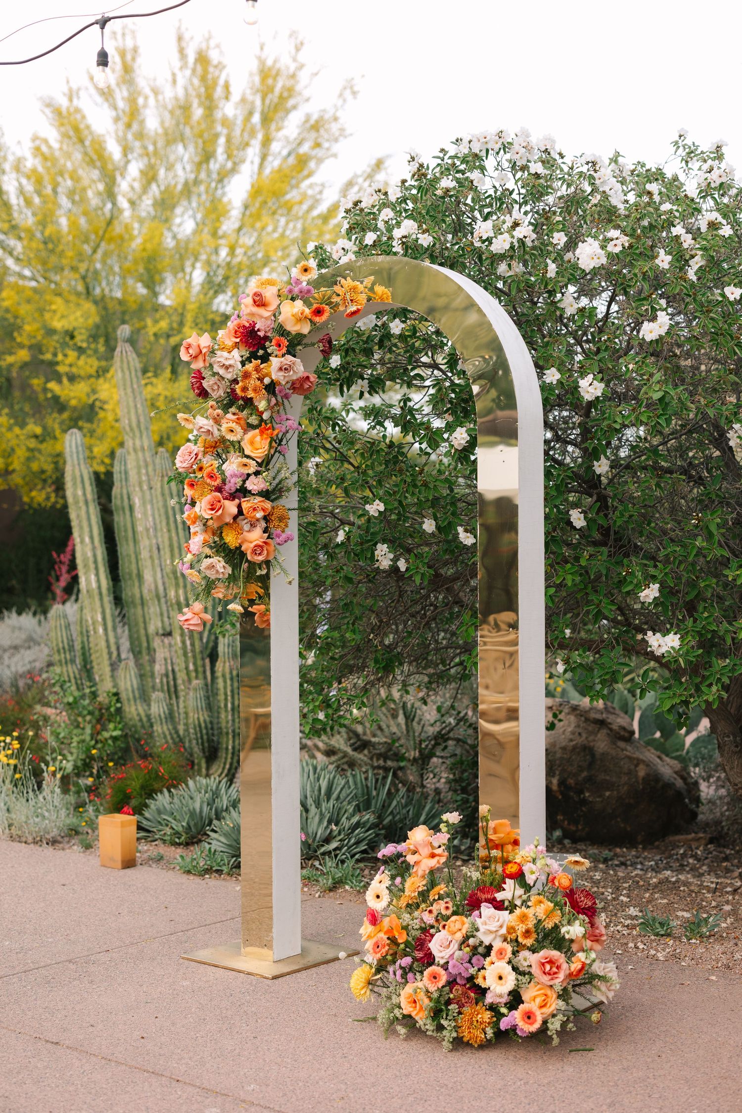 Desert wedding arch decorated with orange and peach flowers alongside towering cacti.