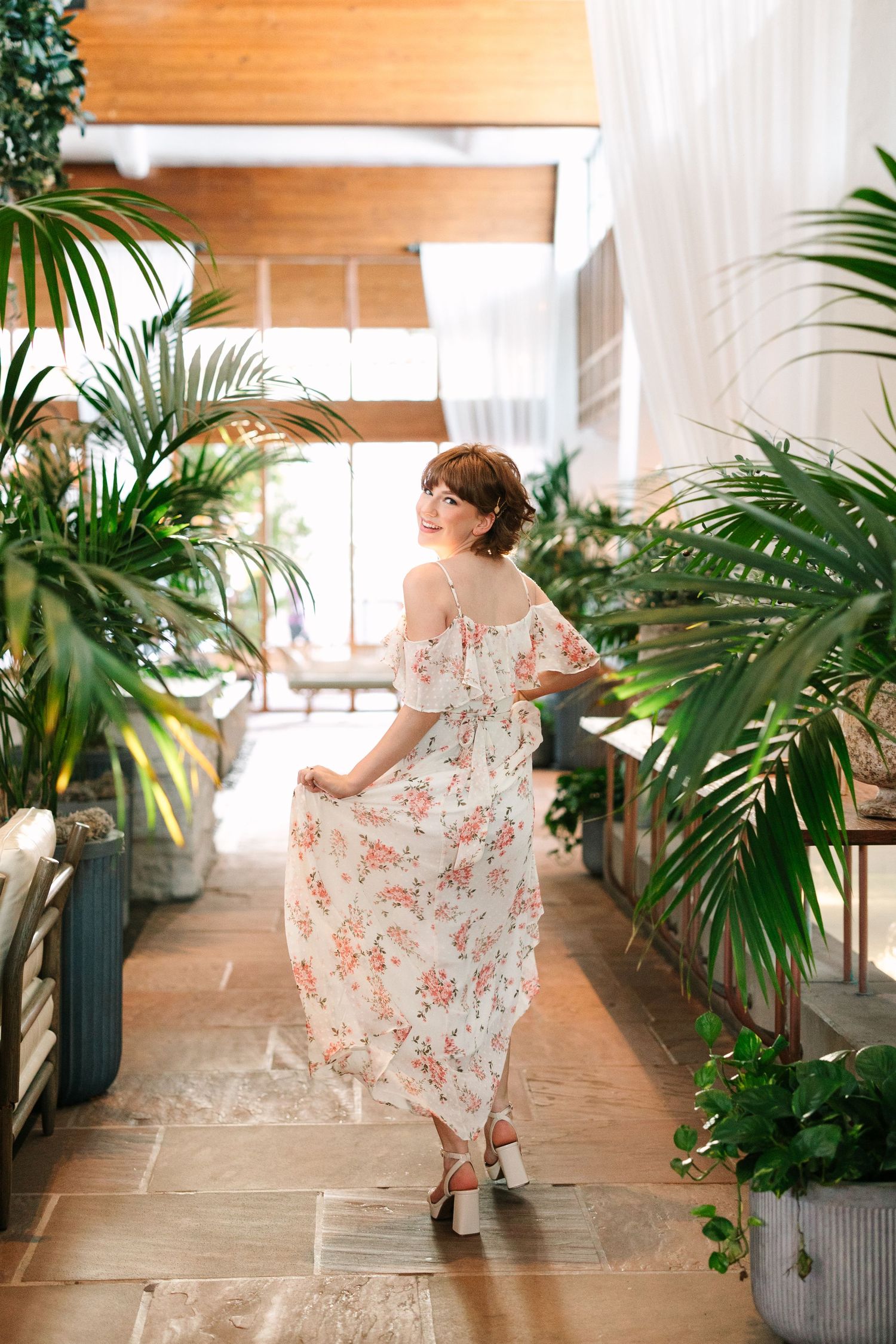 A woman in a floral summer dress twirls gracefully in a bright greenhouse hallway surrounded by tropical palm plants.
