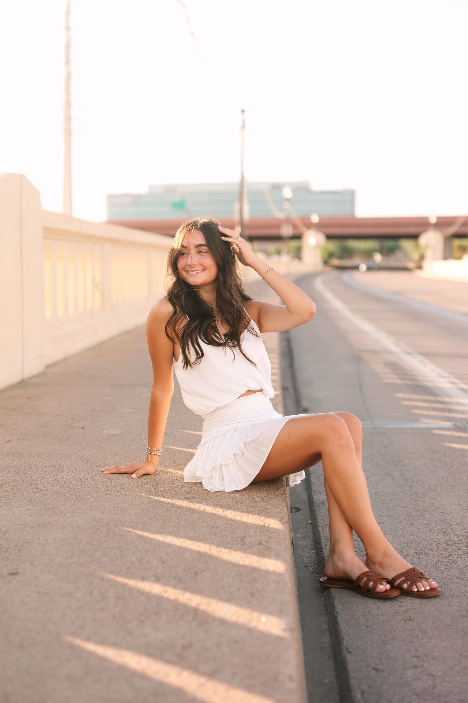 Woman in white dress sits casually on concrete barrier during golden sunset light casting long shadows.