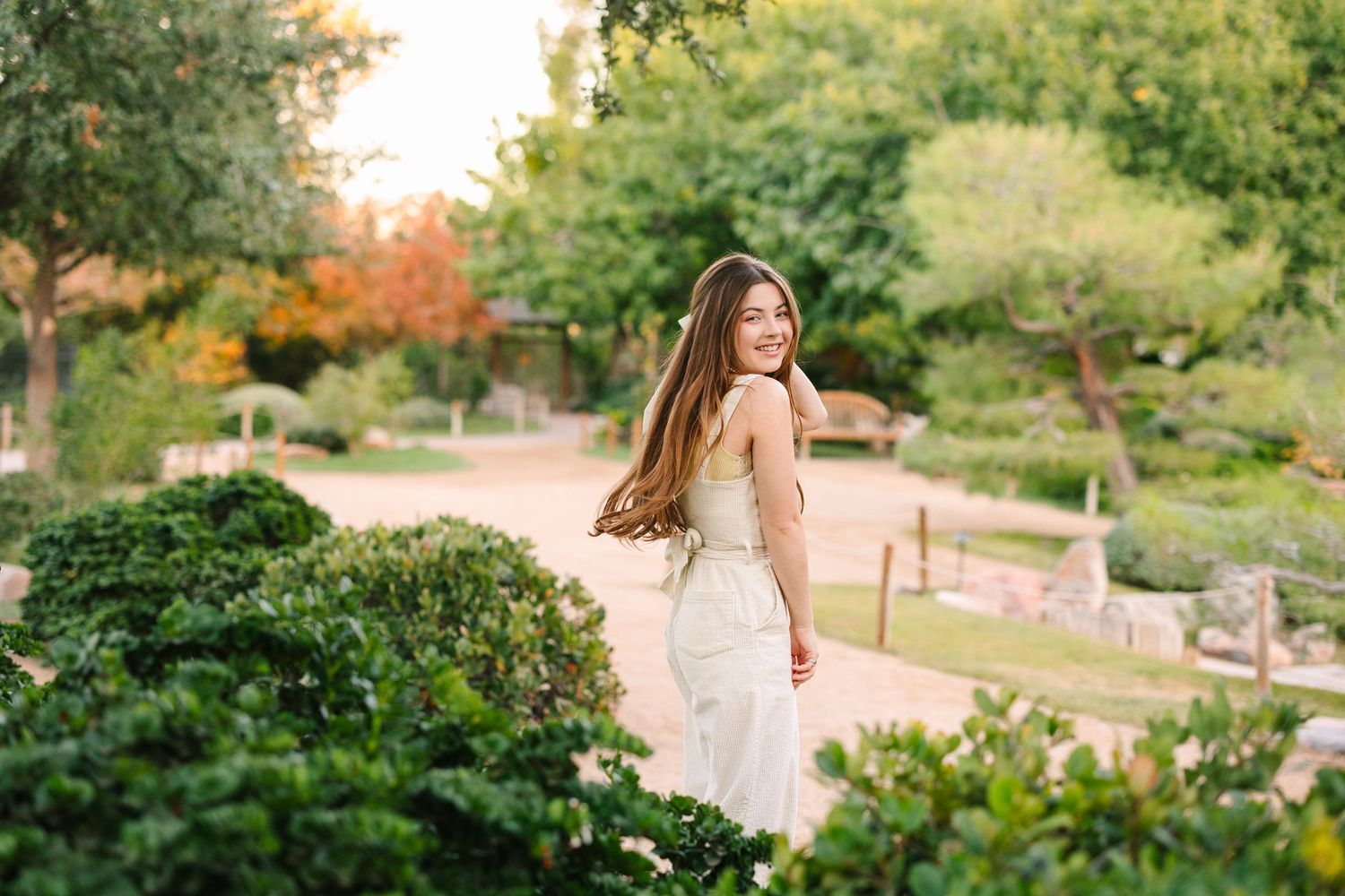 A person in a white gown stands in a lush garden setting during golden hour lighting.