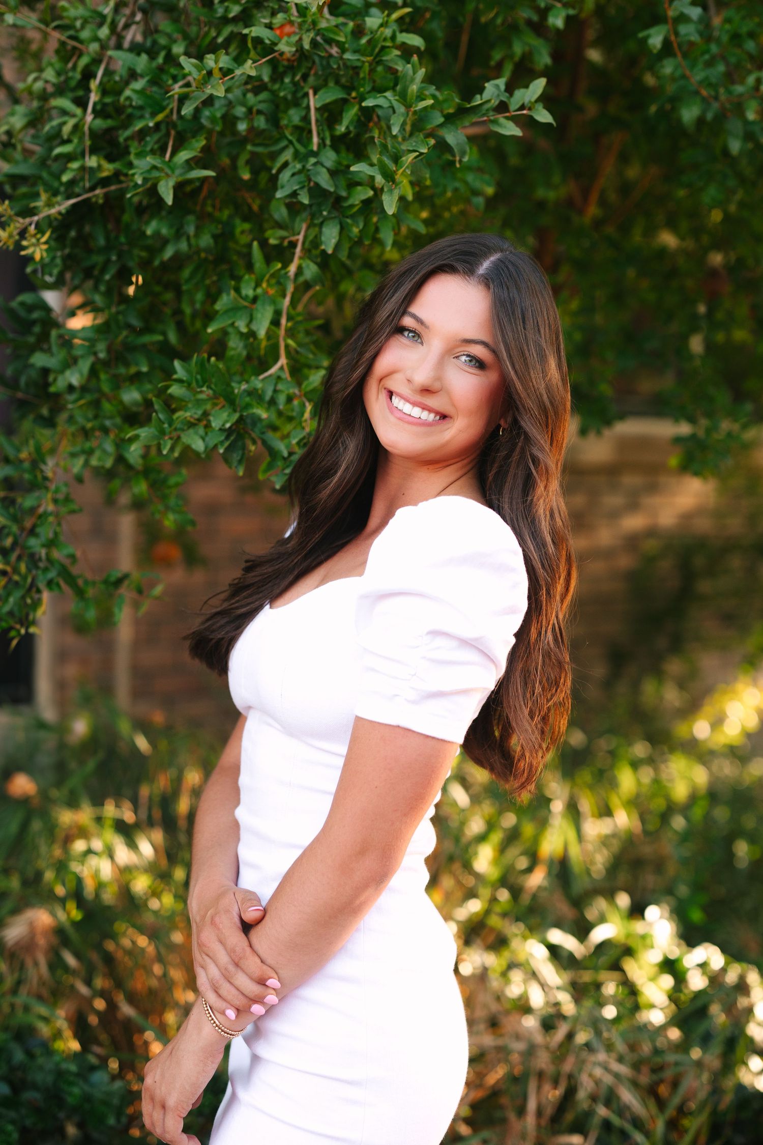 Portrait of a smiling person in a white dress standing outdoors against a green garden backdrop with sunlit foliage.