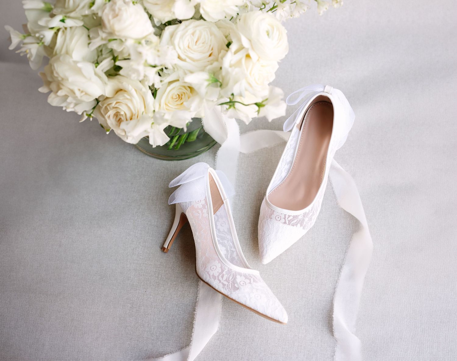 Delicate white bridal shoes with lace details arranged next to white roses. Wedding Flatlay. Molly McPhoto