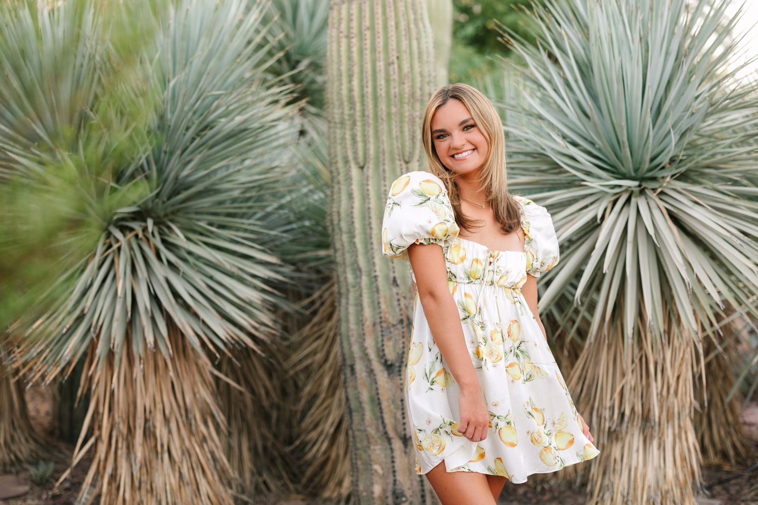 Woman in floral dress poses with spiky desert plants and yucca trees in outdoor garden setting.