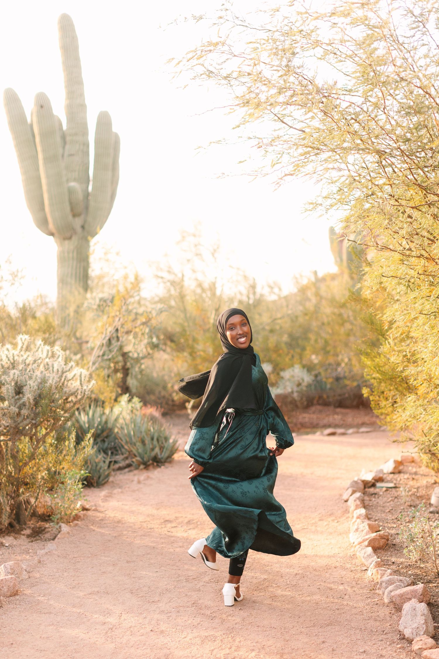 A person in a flowing emerald green dress twirls on a desert path with a majestic saguaro cactus in the background.