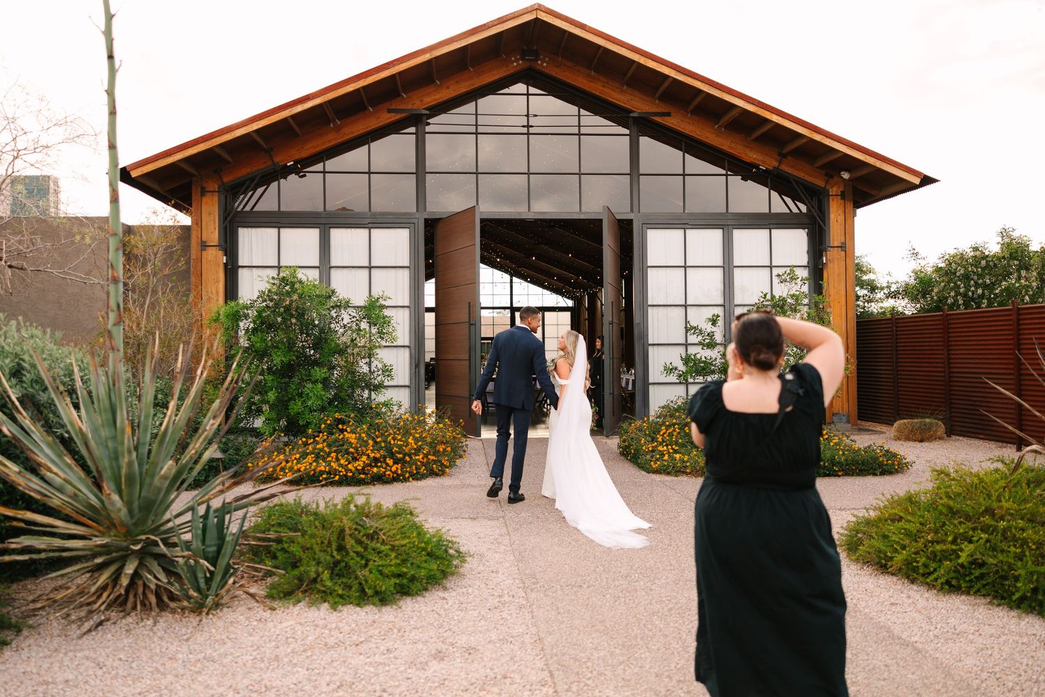 Photographer capturing couple entering modern glass-front wedding venue with desert landscaping. The Paseo, Apache Junction AZ
