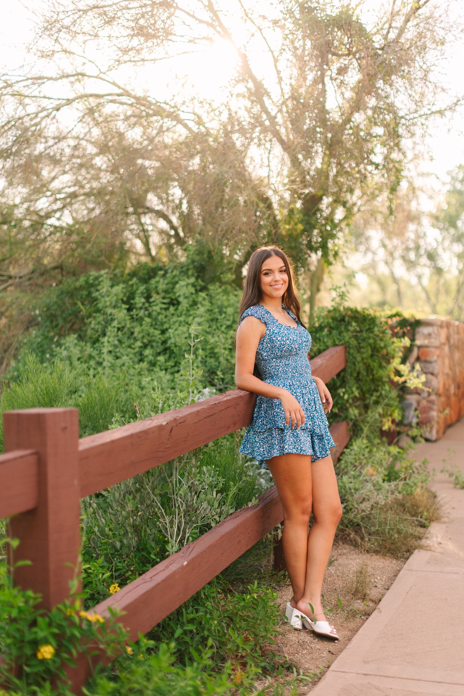 A blue floral sundress and sandals outfit photographed against a rustic wooden fence and natural greenery at sunset.