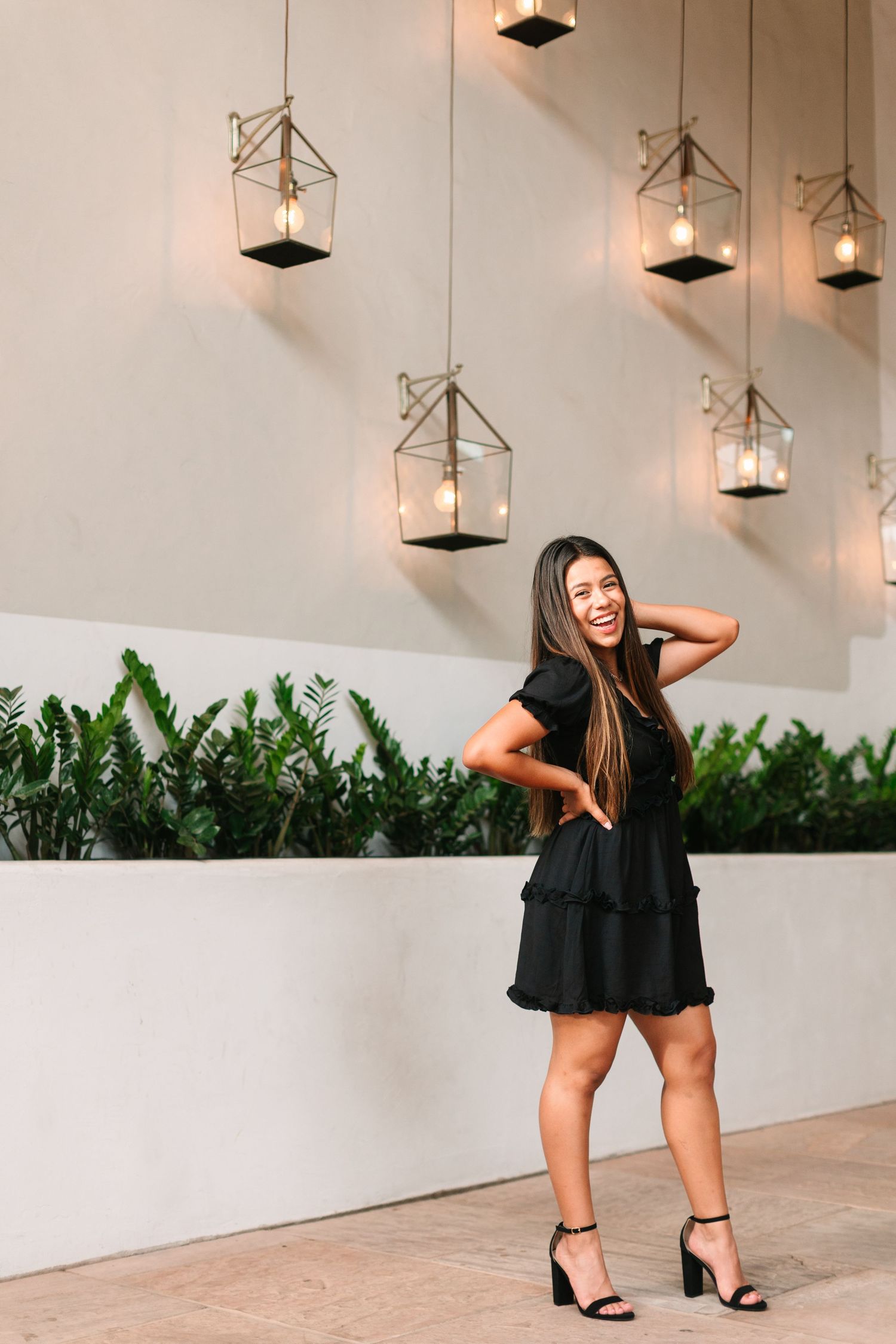A person in a black dress poses playfully against a white wall with hanging pendant lights and green plants.