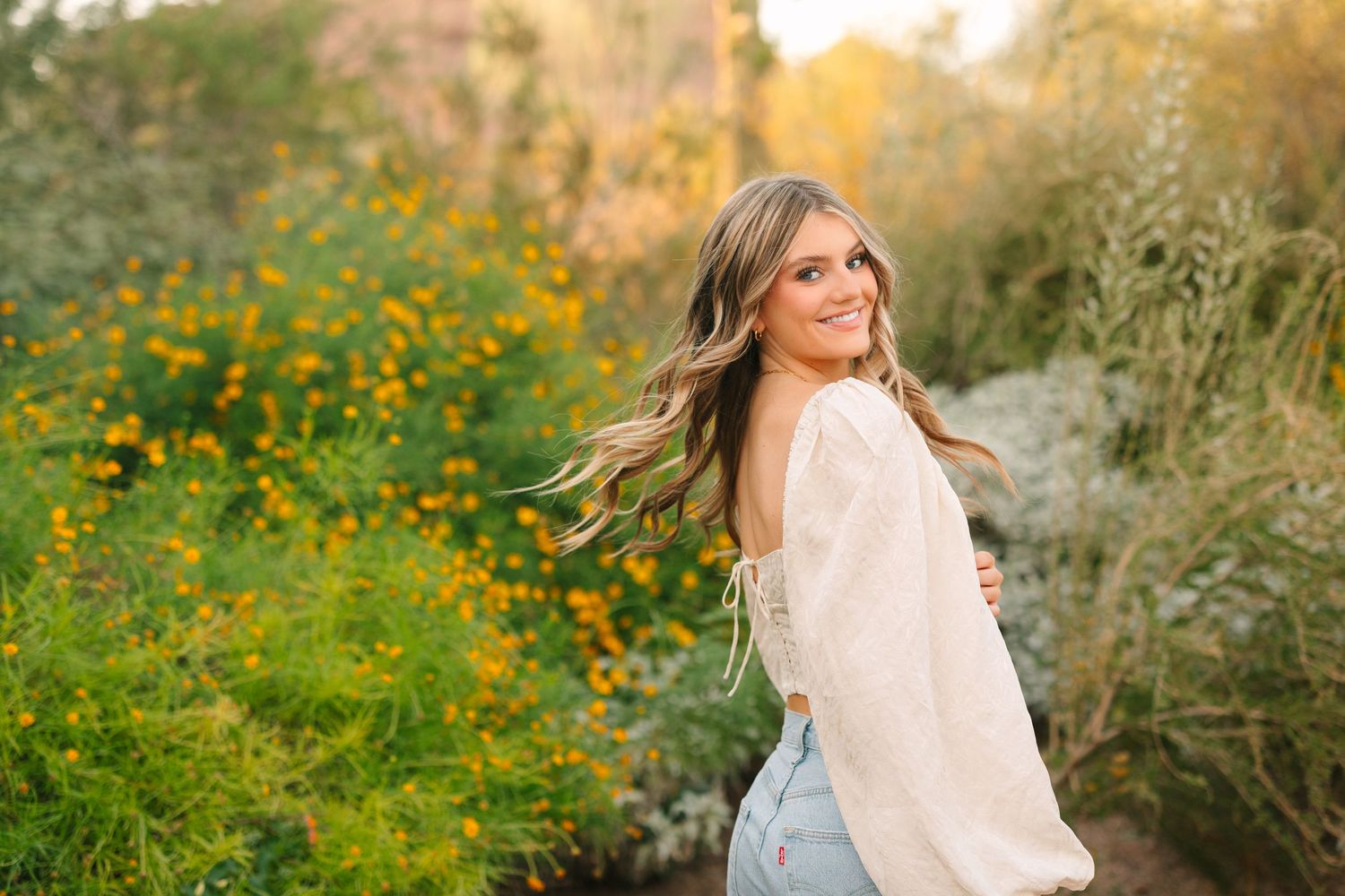 Woman in white lace dress poses amidst golden wildflowers in a dreamy outdoor setting during sunset.