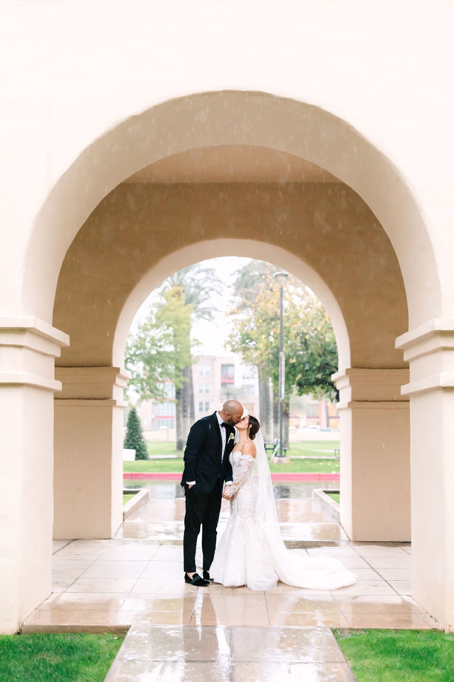 Wedding couple poses under stone archway on rainy day.