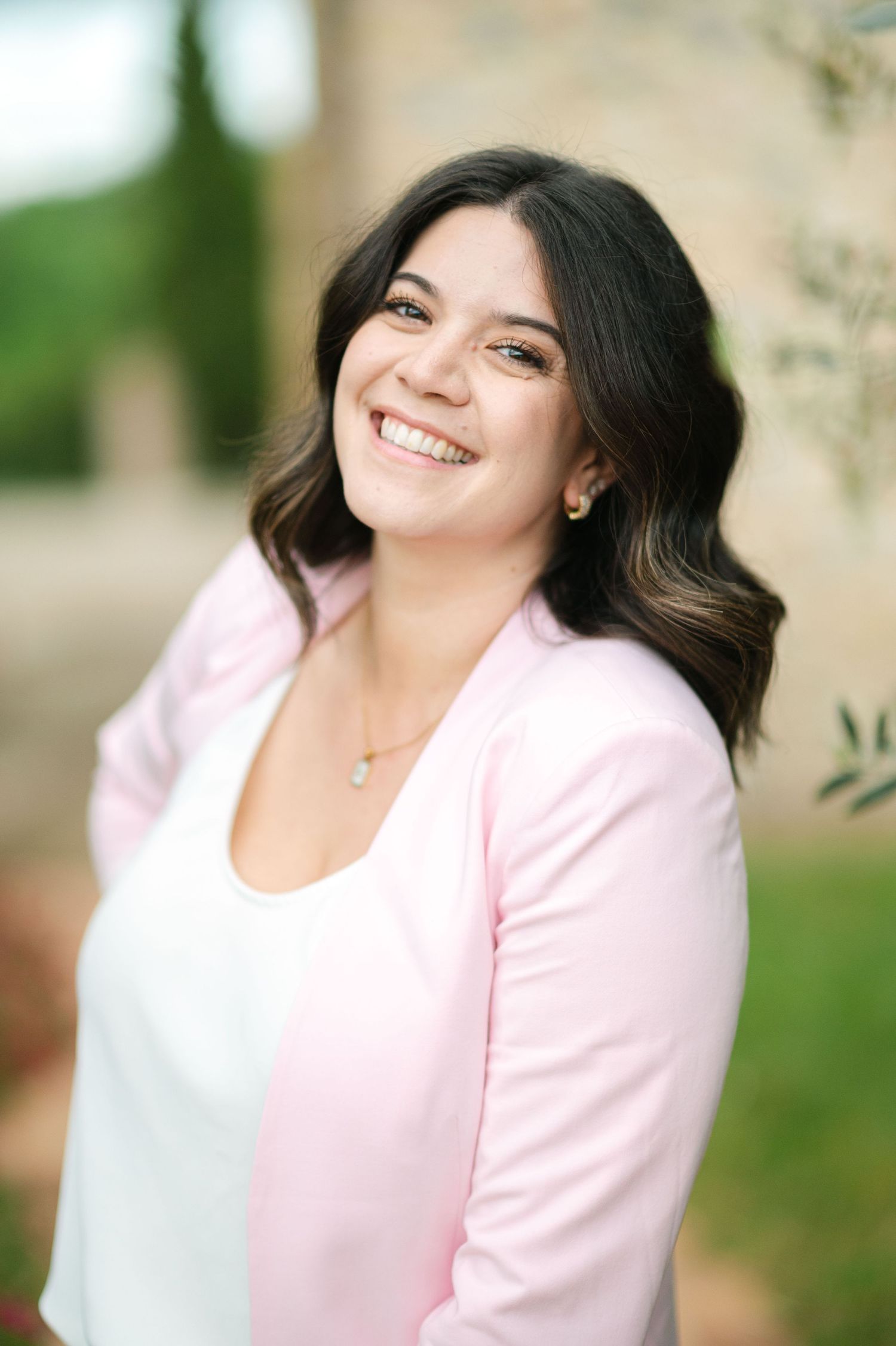 A woman in a light pink cardigan and white top smiles warmly while posing outdoors with greenery in the background.