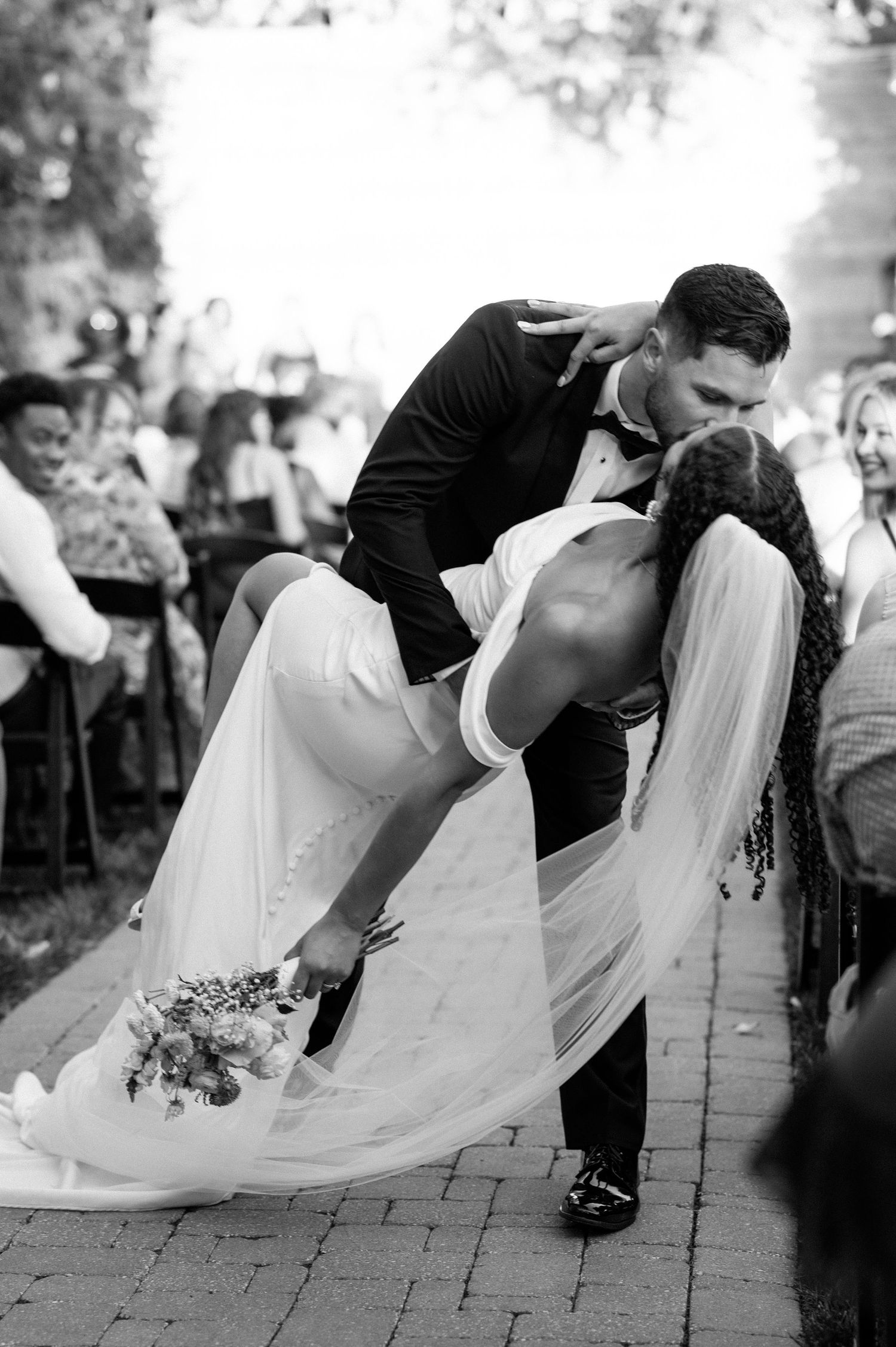 A dramatic dip kiss between newlyweds in black and white as guests look on during an outdoor wedding celebration.