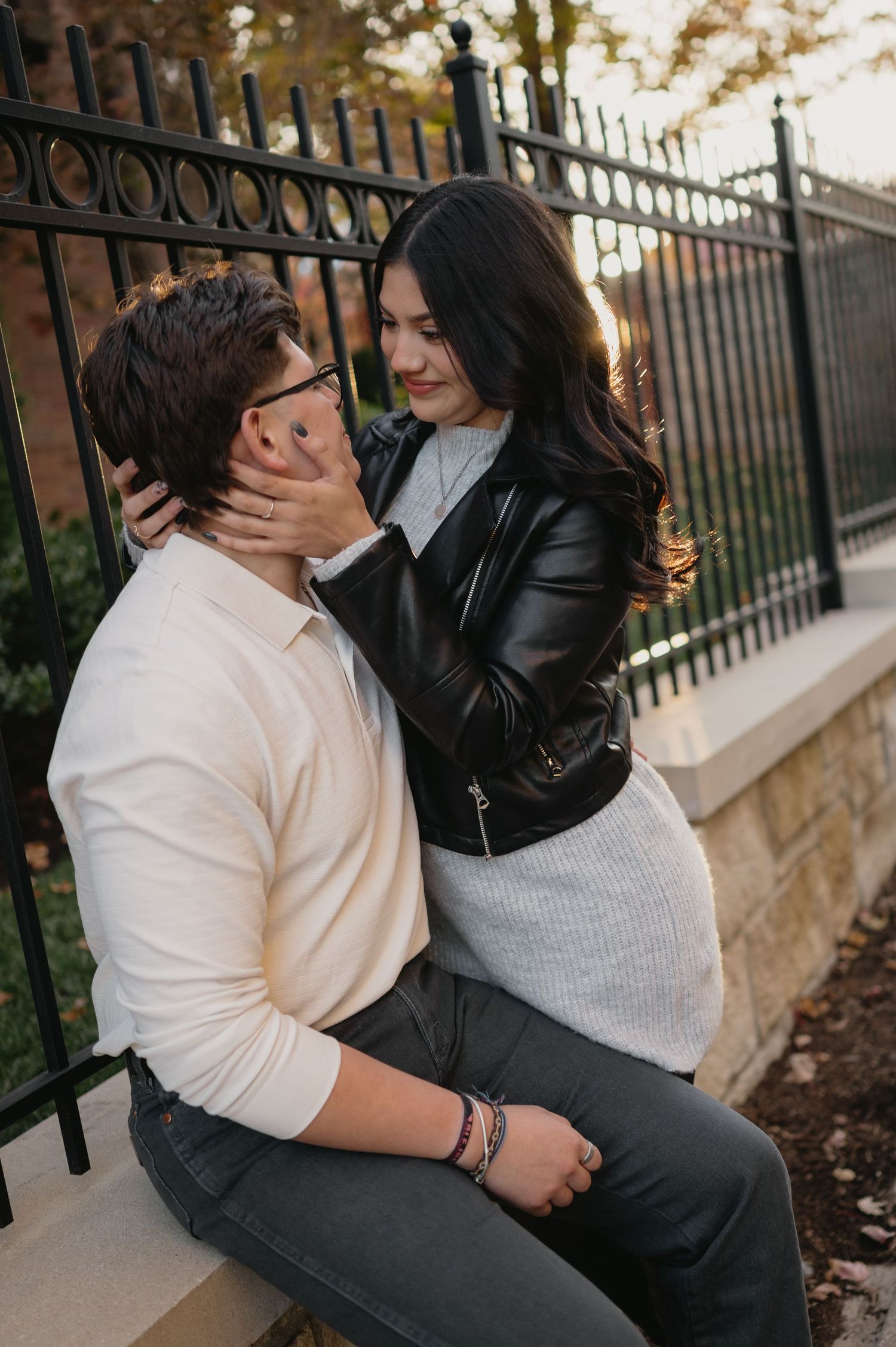 Romantic couple embracing by an iron fence at sunset with person in leather jacket holding their partner tenderly.