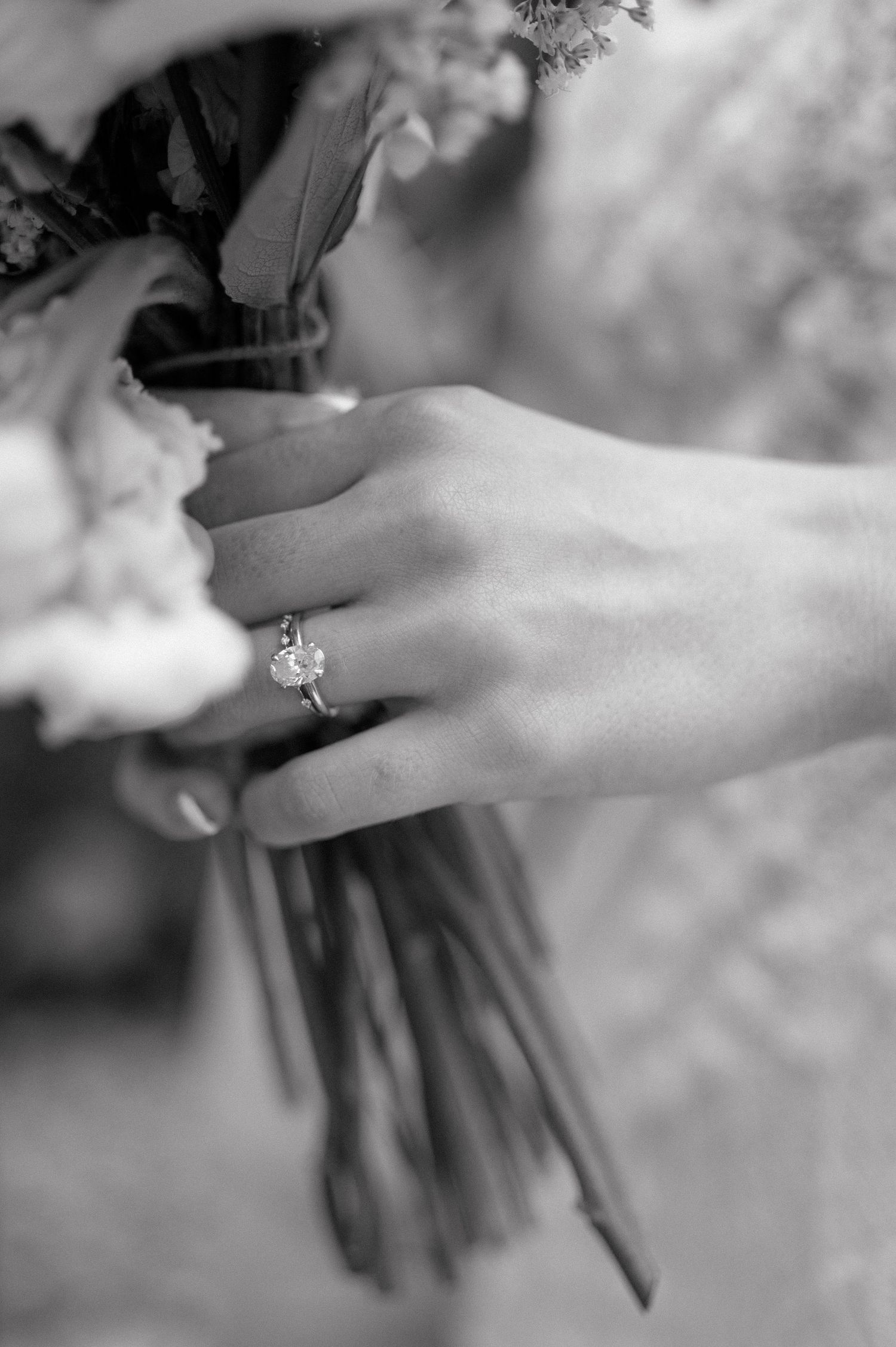 Black and white close-up image of a hand wearing an elegant diamond engagement ring while holding a bouquet of flowers.