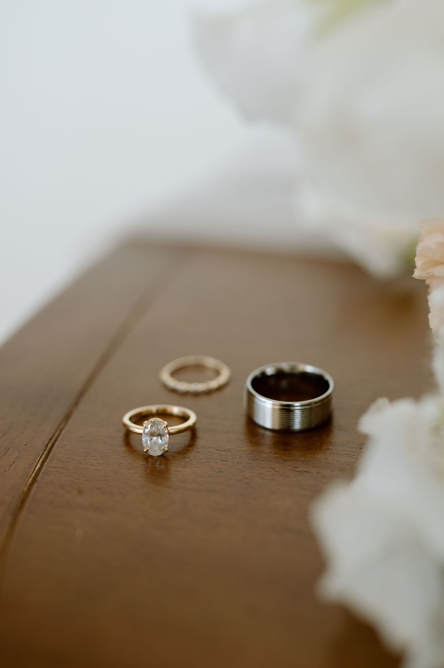 Wedding rings and engagement ring with diamond solitaire arranged on wooden surface with white floral accents.