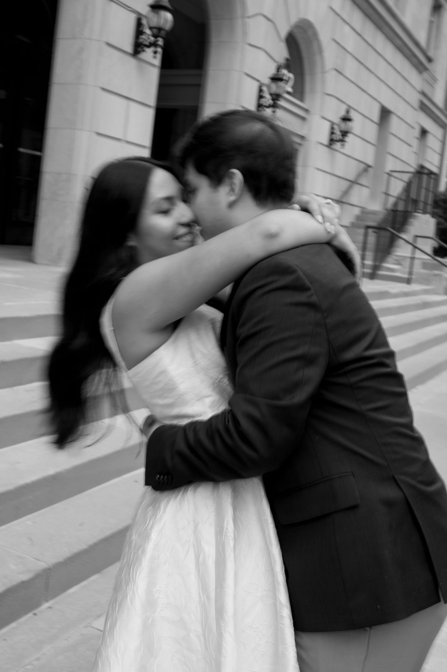 A romantic black and white photograph of a couple embracing and kissing on outdoor steps by a building with wall lamps.