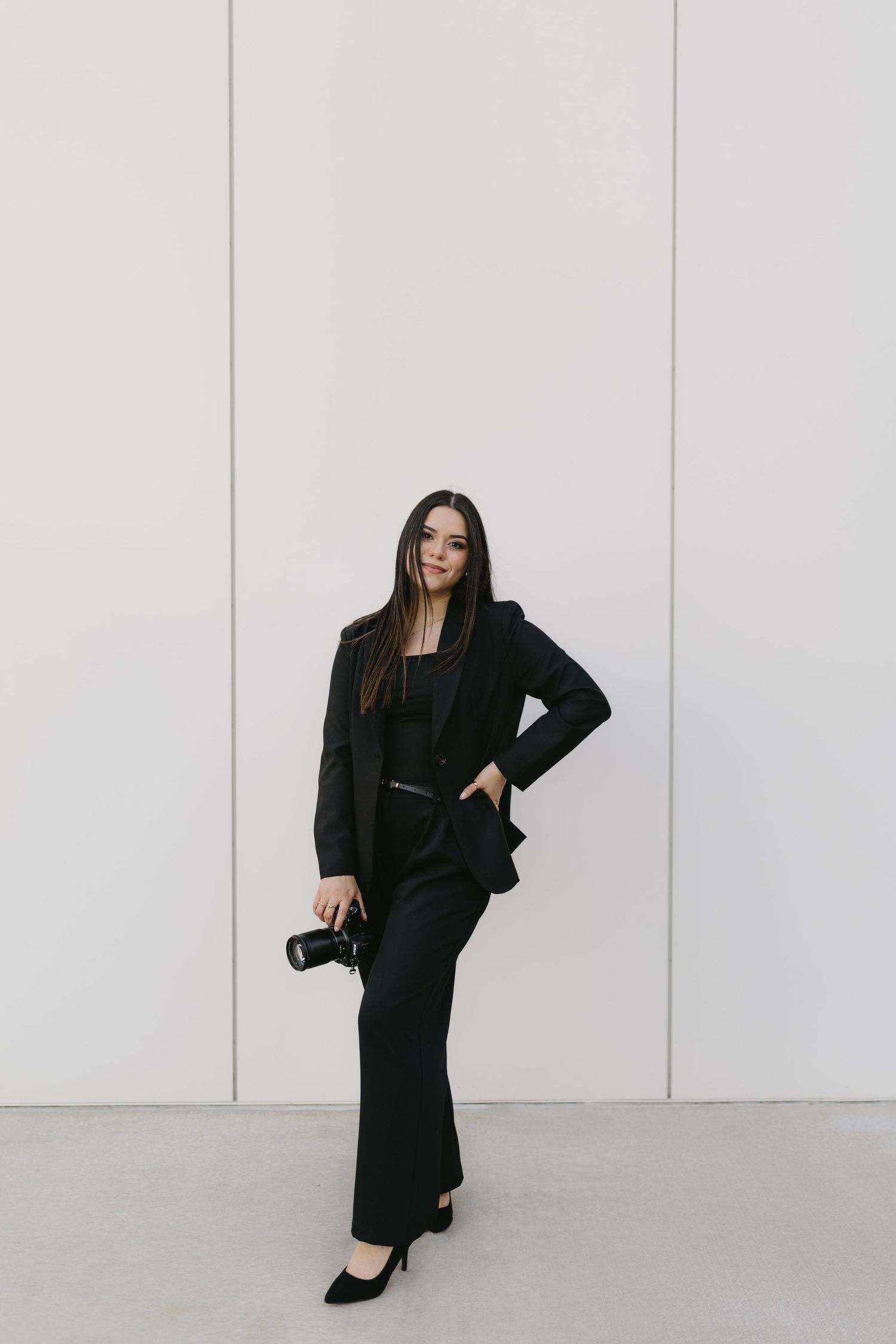 Professional photographer in an all-black outfit stands against white wall holding camera with confident pose.