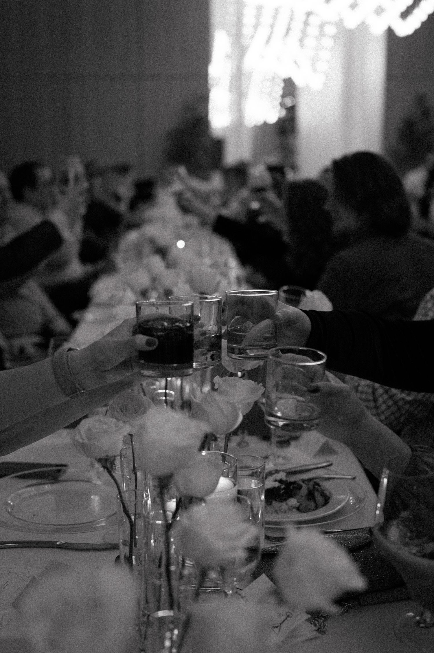 Black and white photo of a wedding reception dinner table with wine glasses, plates and dining guests in soft focus.