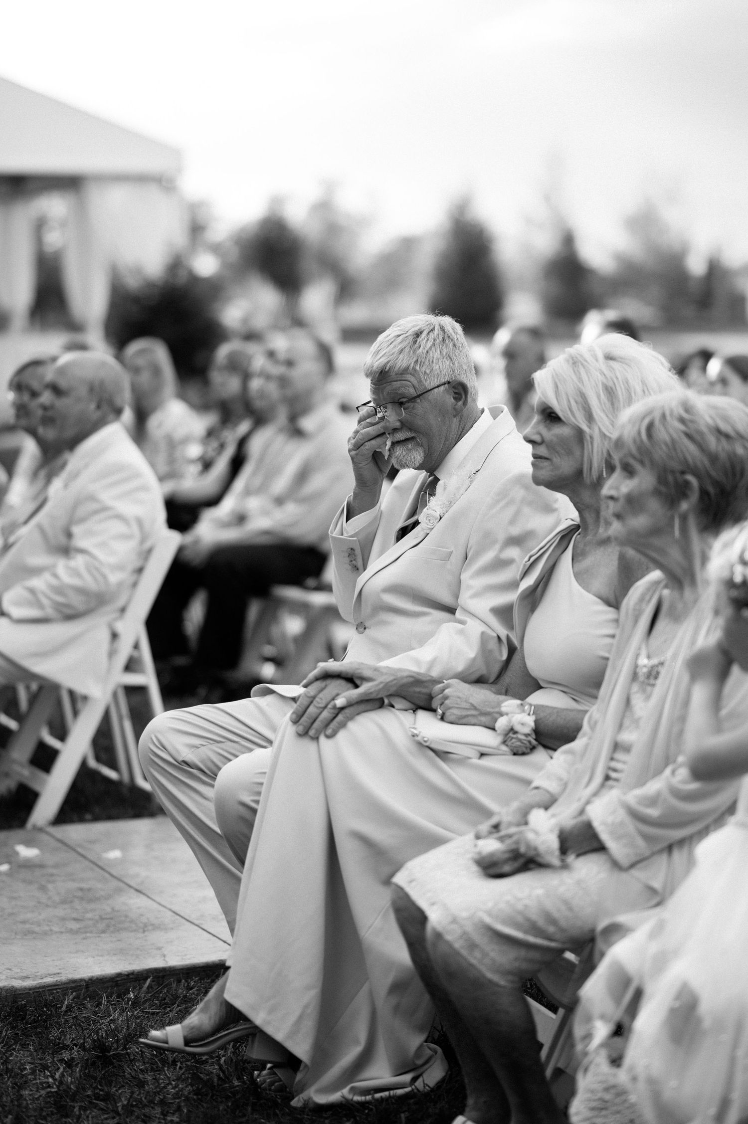 Black and white photograph shows crowd seated in white folding chairs at outdoor event, with attendees wearing light colored clothing.