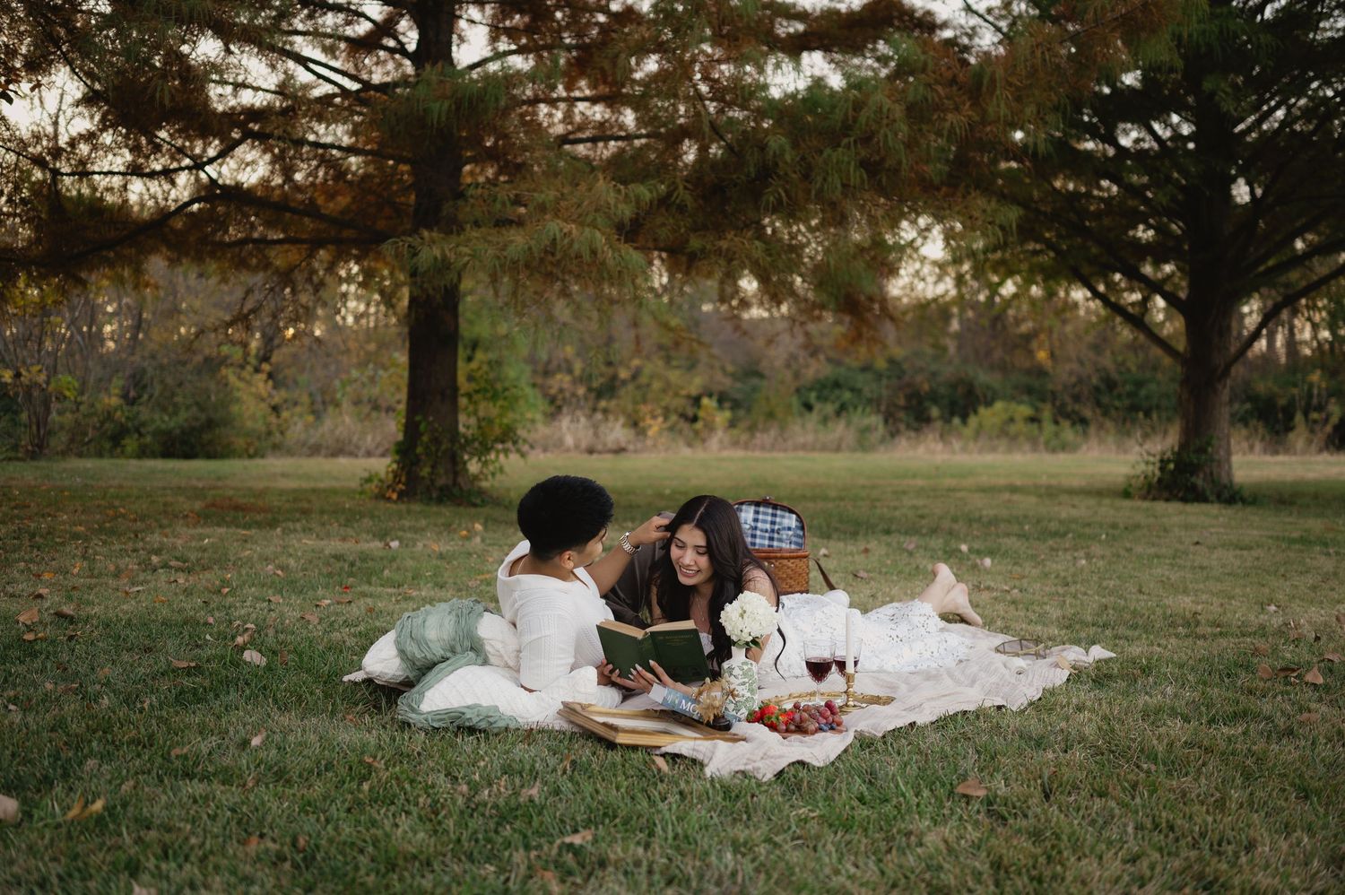 Three people enjoying a cozy picnic on a blanket in a grassy park under tall pine trees at golden hour.