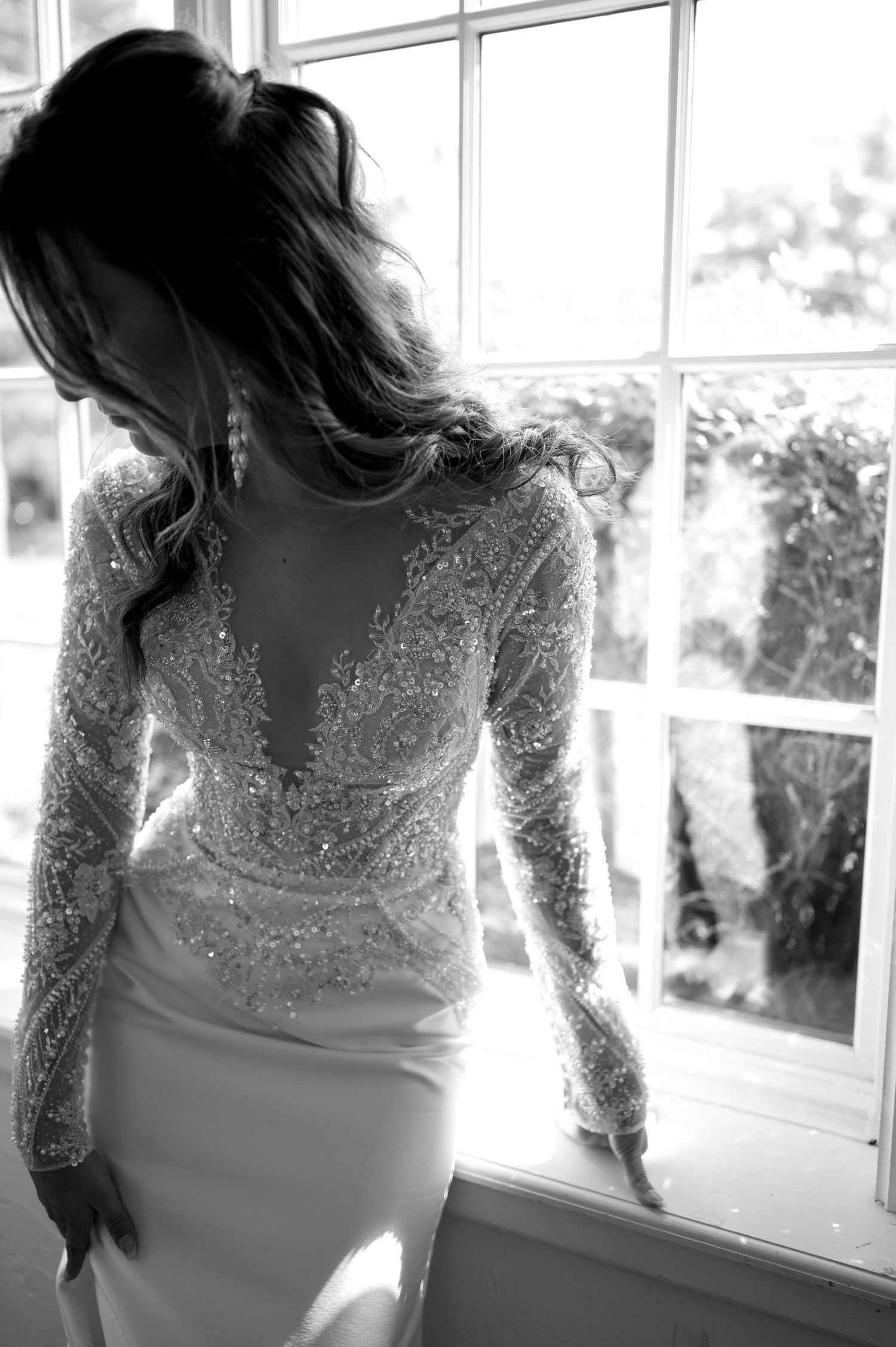 A backlit silhouette of a bride wearing a fitted lace wedding gown with intricate long sleeves stands by a bright window.