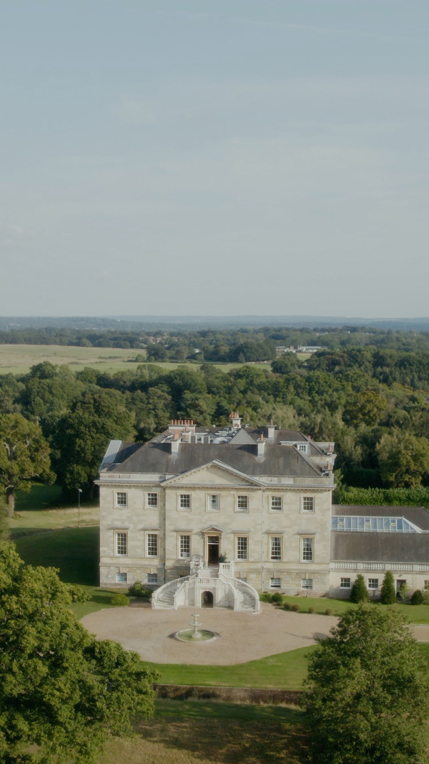 Elegant Georgian manor house with grand entrance steps surrounded by lush countryside and gardens in rural setting.