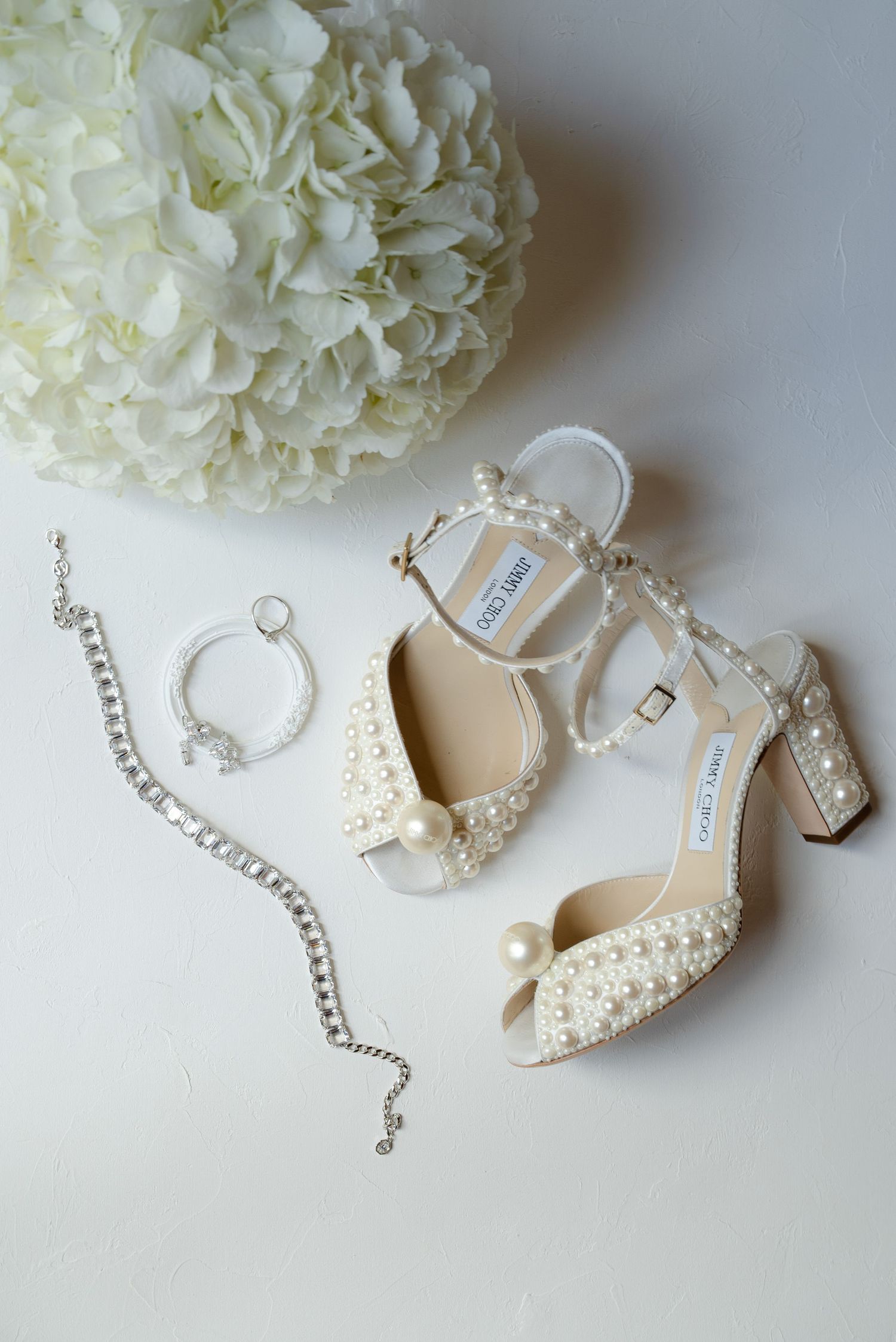 Elegant bridal accessories including pearl-studded heels, silver jewelry, and a white flower arrangement on light background.