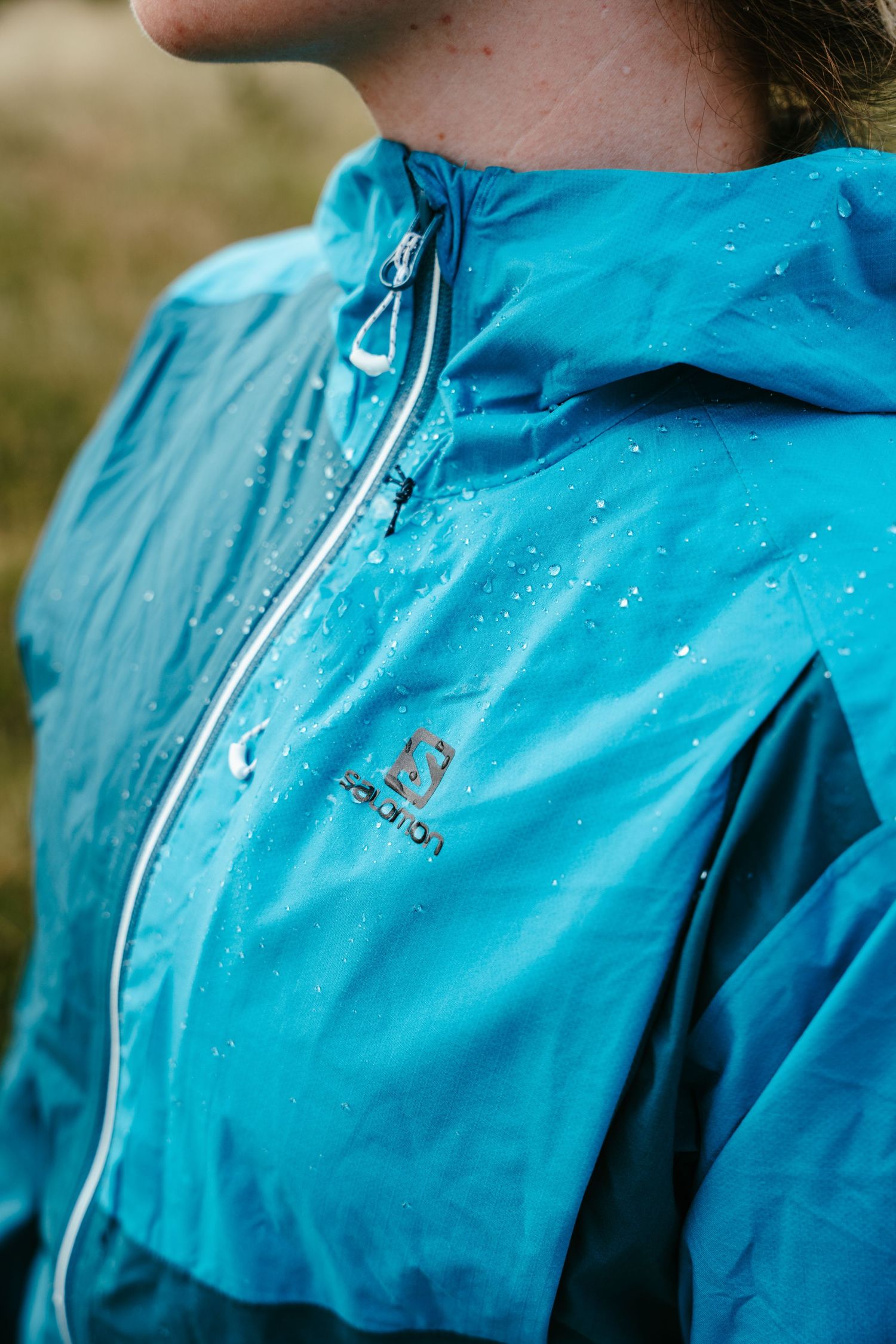 Closeup of a turquoise blue waterproof jacket with water droplets and the Salomon logo visible on the fabric.