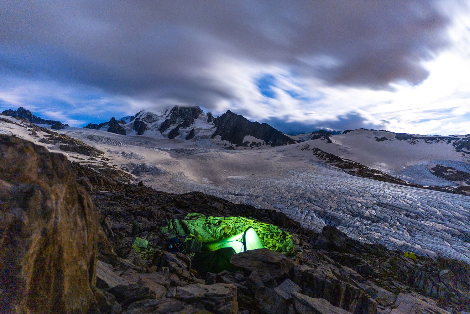 Illuminated green camping tent glows against dramatic mountain landscape with glaciers and stormy clouds at dusk.